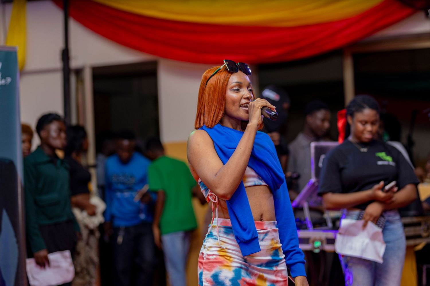 Performer with bright orange hair sings on stage with decorative banners in background.