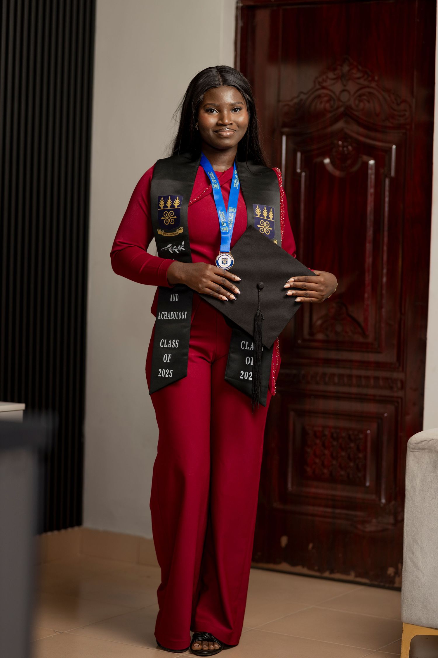 Young woman in red jumpsuit holding graduation cap, wearing black stole and medal, posing proudly indoors.