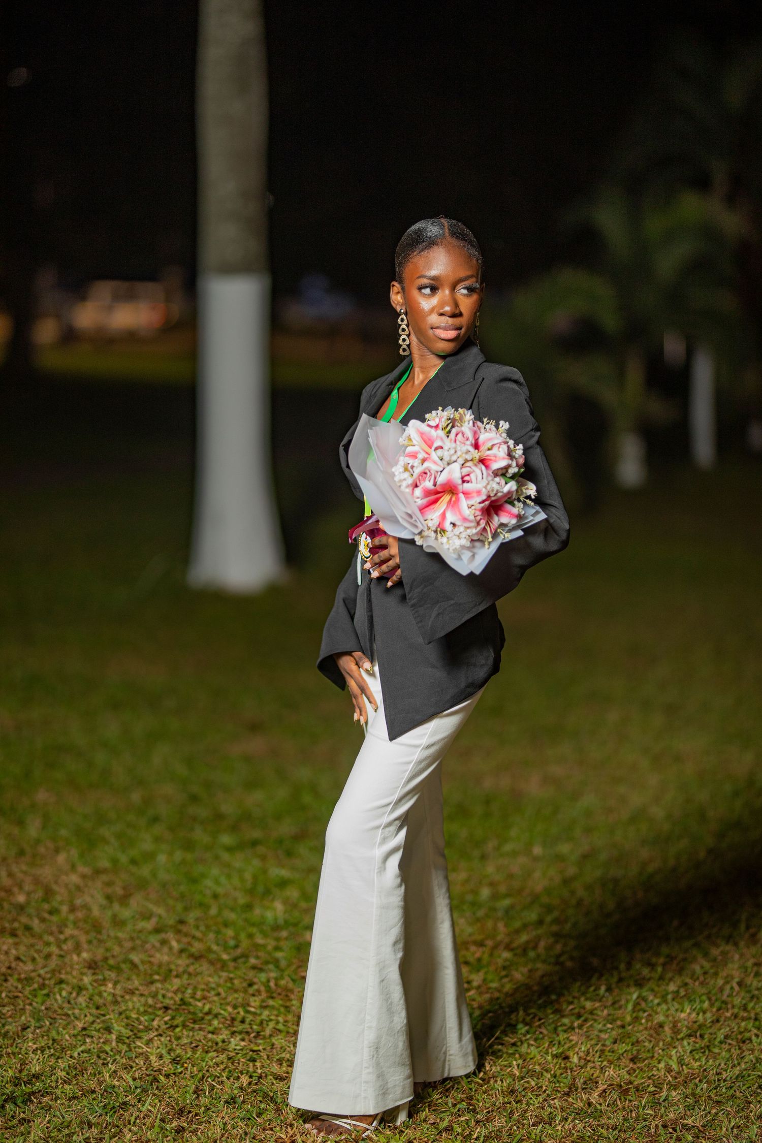 A person in a white dress and black jacket poses in a grassy outdoor setting at dusk.
