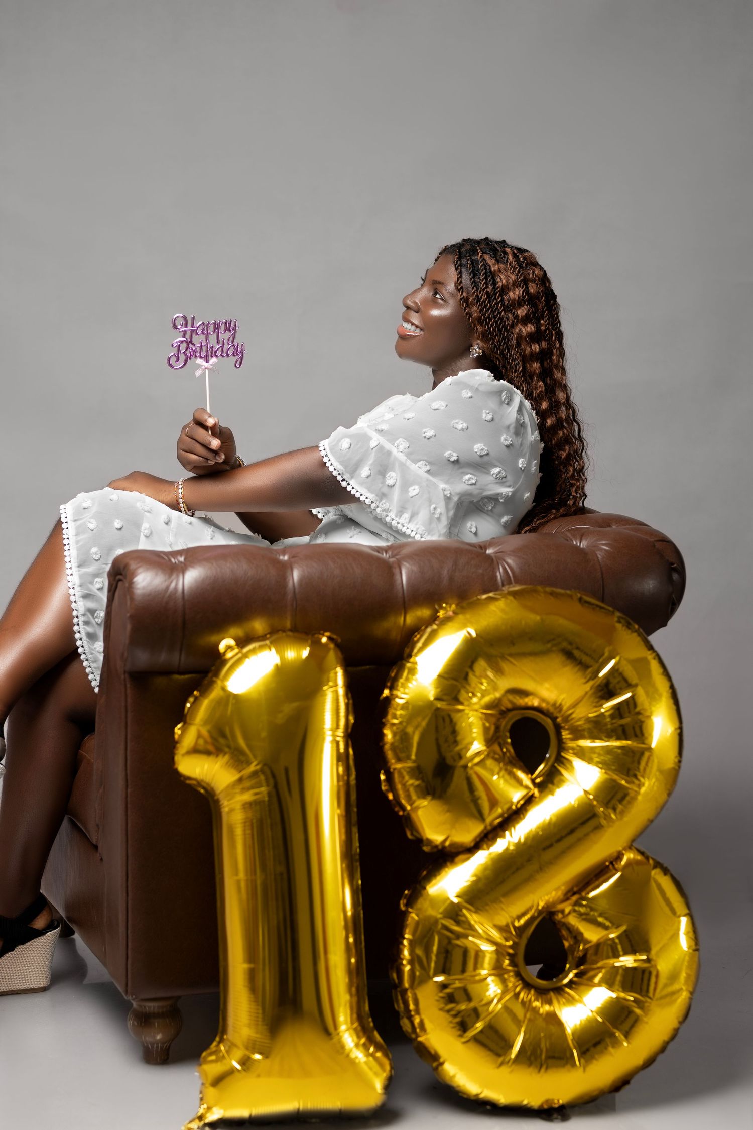 A young woman in white dress reclines against a brown chair with gold '18' balloons for a birthday photo.