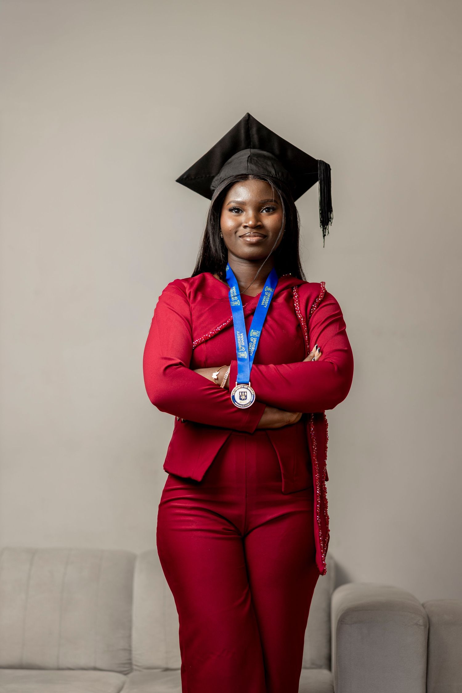 A young woman in a red suit and graduation cap poses confidently with arms crossed, wearing a blue medal.