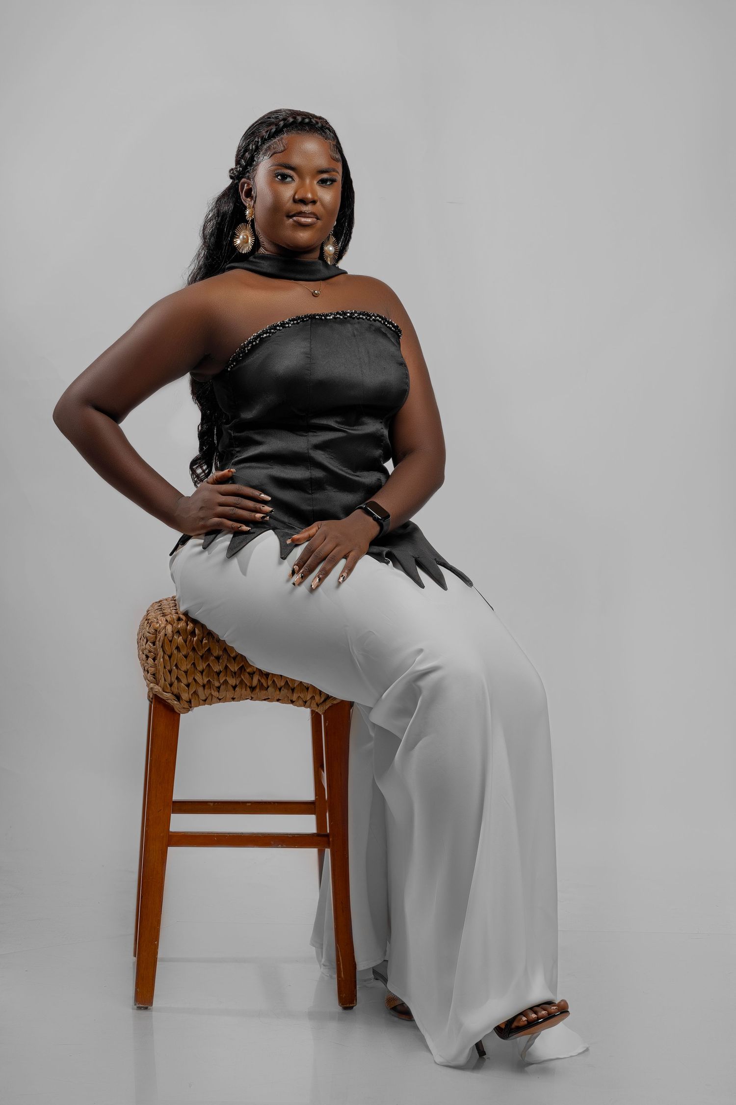 Stylish plus-size fashion photoshoot featuring a black top and white pants ensemble staged on a wooden stool against gray backdrop.