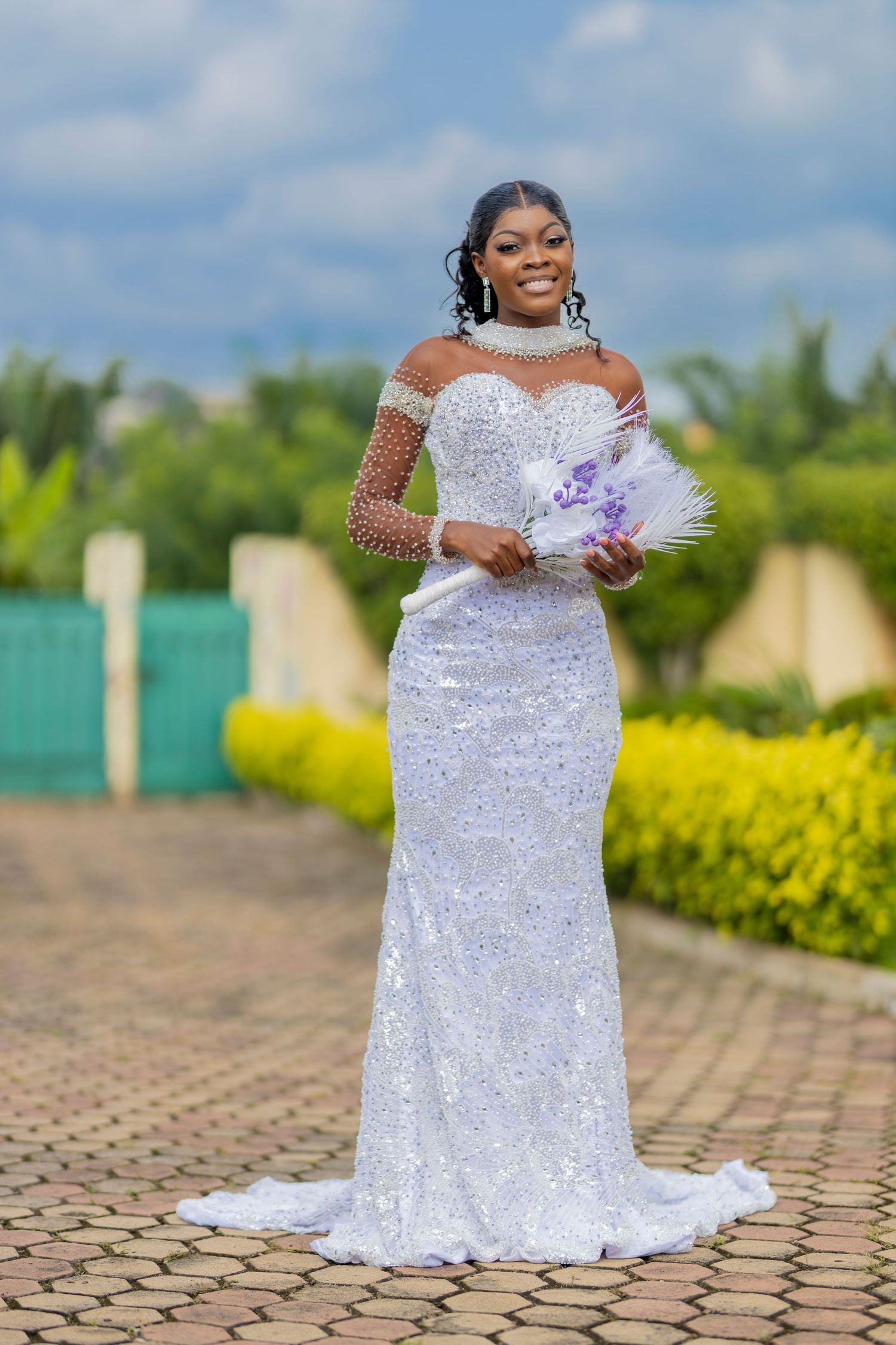 A stunning bride in a sparkling white wedding dress poses outdoors in a garden setting with stone pavers and greenery.