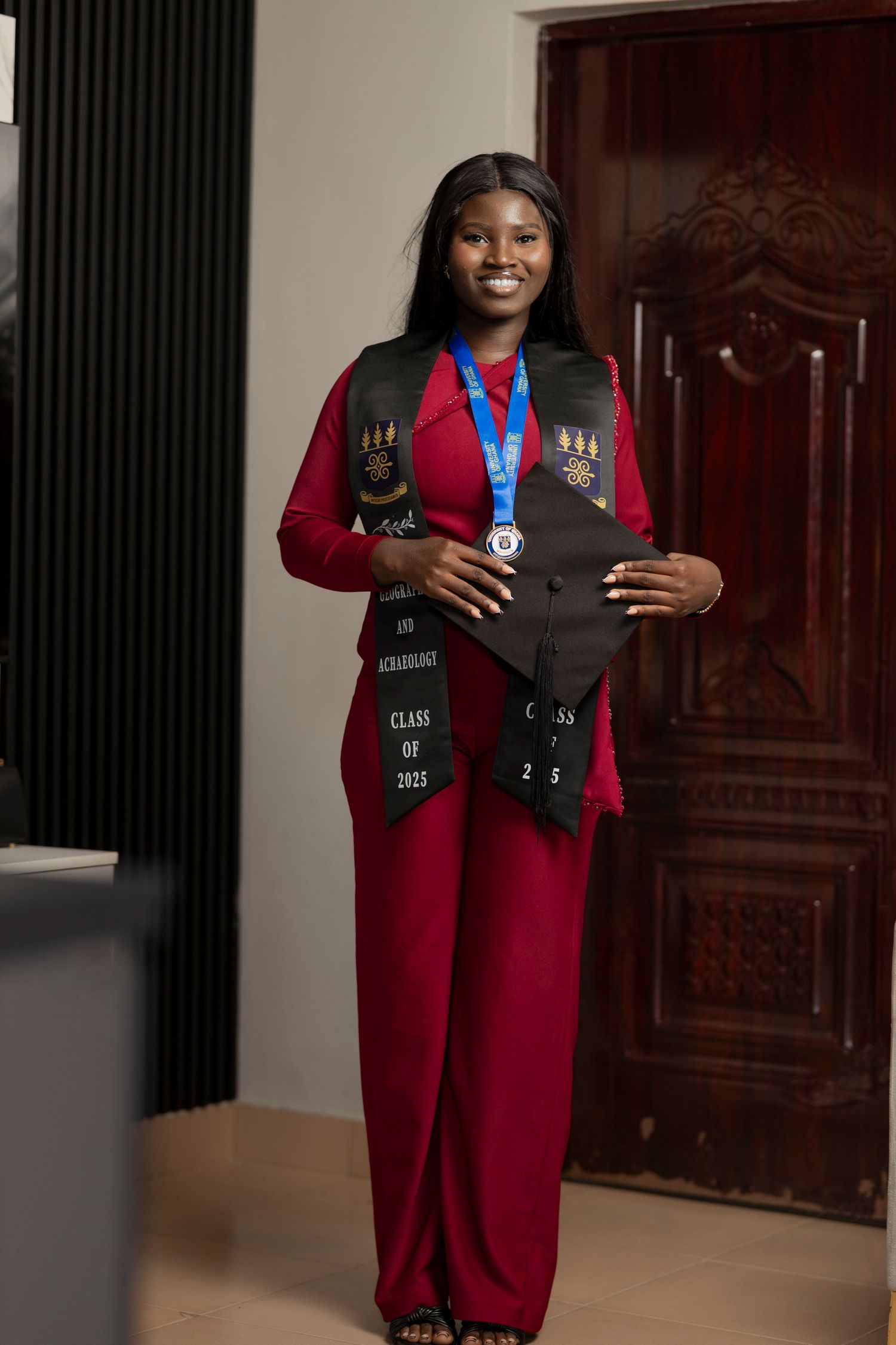 Smiling graduate in red outfit holding cap with black stoles and medal, Class of 2025 graduation photo.