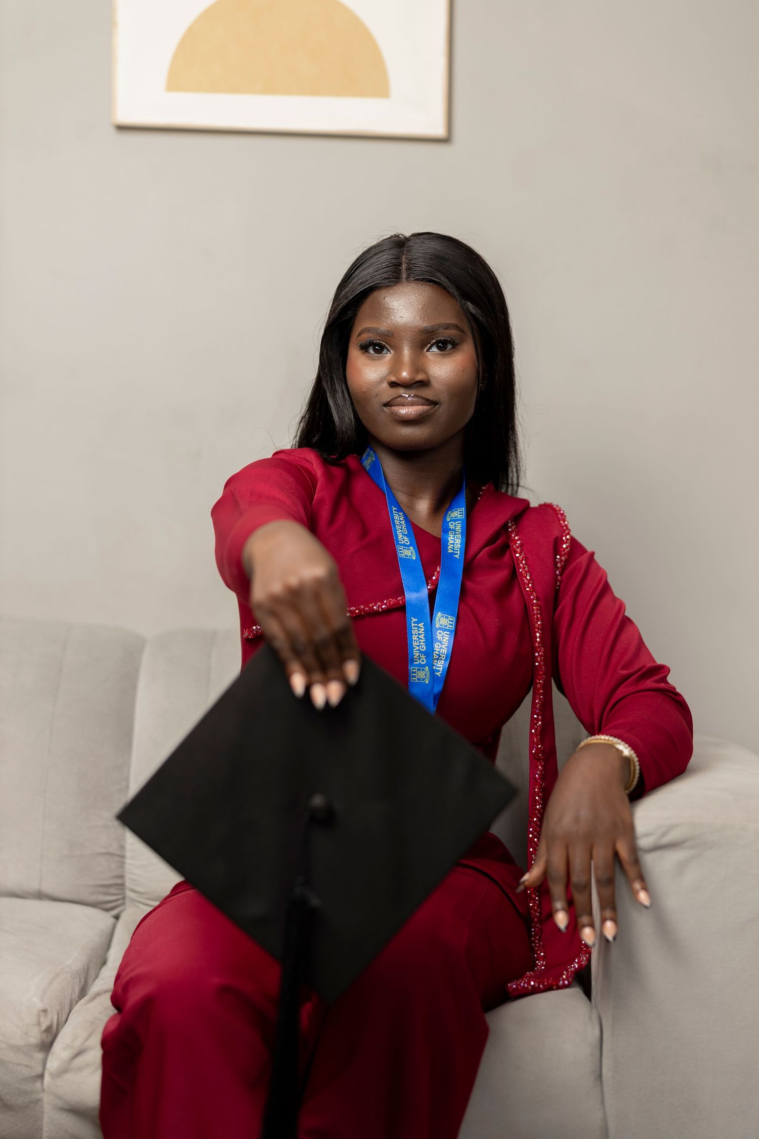Young Black woman in red outfit holding graduation cap with blue medal, sitting on couch indoors.