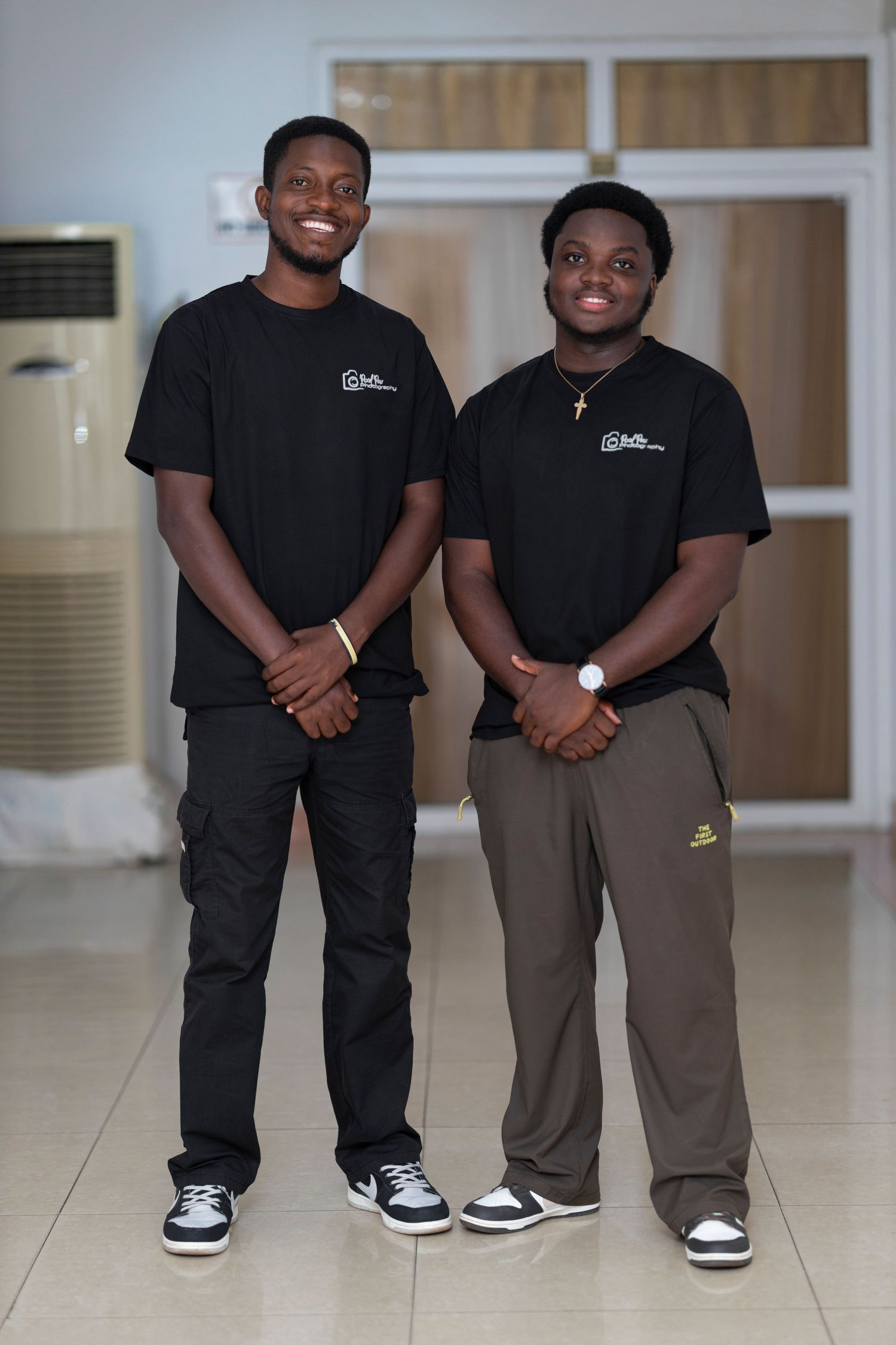 Two young men wearing matching black t-shirts and casual pants smile while posing together indoors.