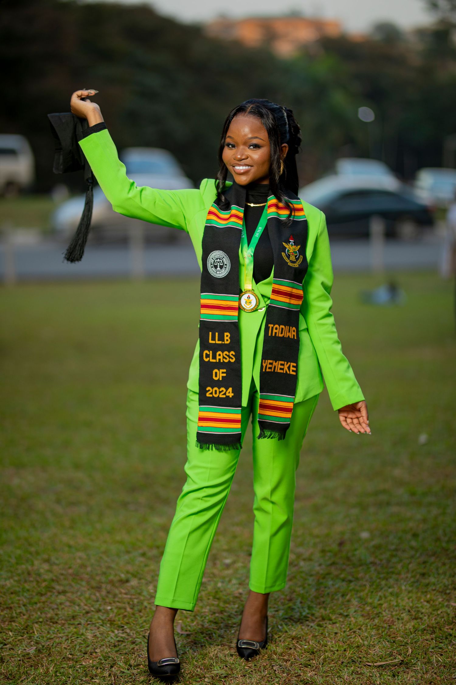 Graduate wearing bright lime green suit and cultural stole poses joyfully on grass field during commencement celebration.