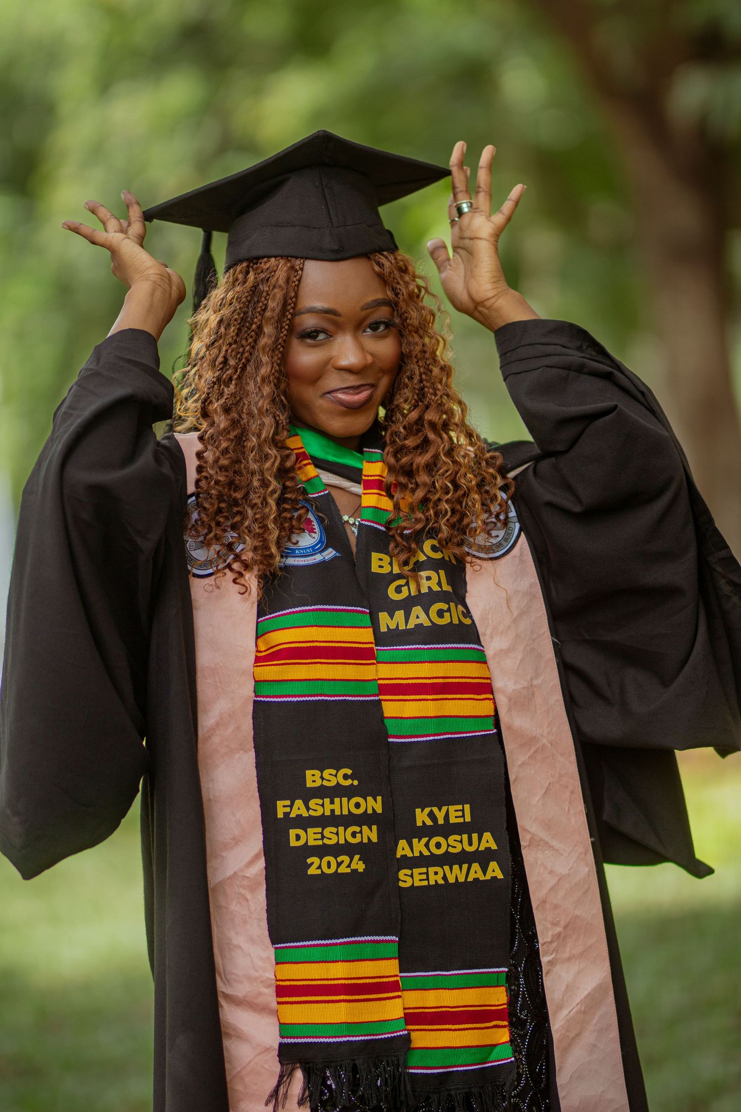 Graduate wearing black cap and gown with colorful kente stole and BSc Fashion Design graduation regalia poses outdoors.