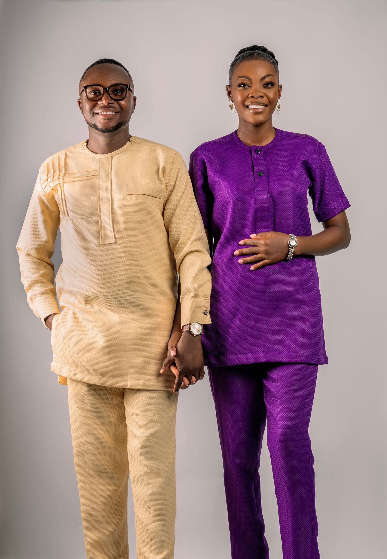 Two people pose together wearing contrasting cream and purple loungewear sets in a studio setting.