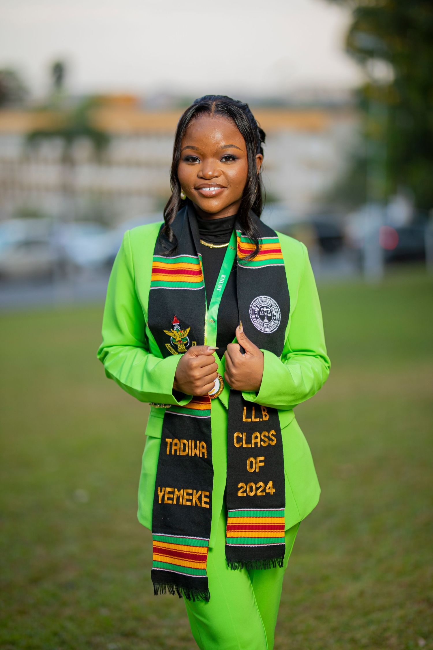 Graduate in bright neon green academic regalia poses with class of 2024 graduation stole in outdoor setting.