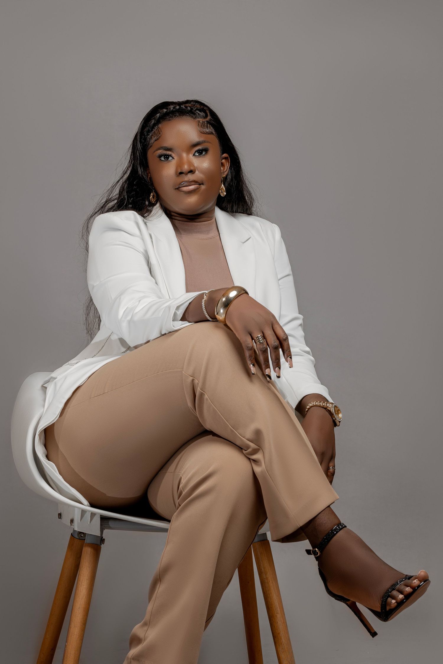 Professional studio portrait showing a model in a white blazer and tan pants seated elegantly on a wooden stool.