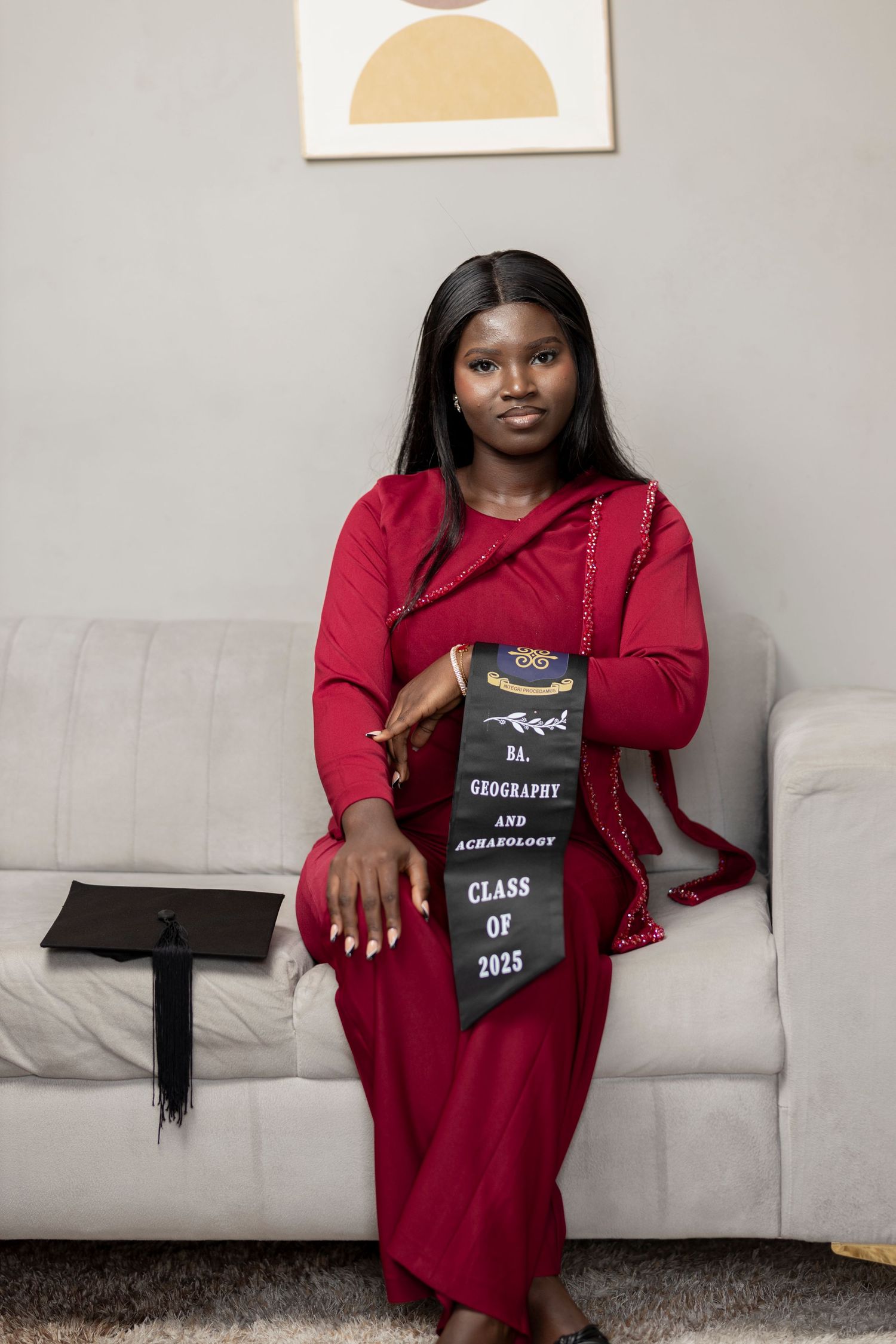 Young woman in red dress wearing Class of 2025 Geography and Archaeology graduation stole seated on white sofa.