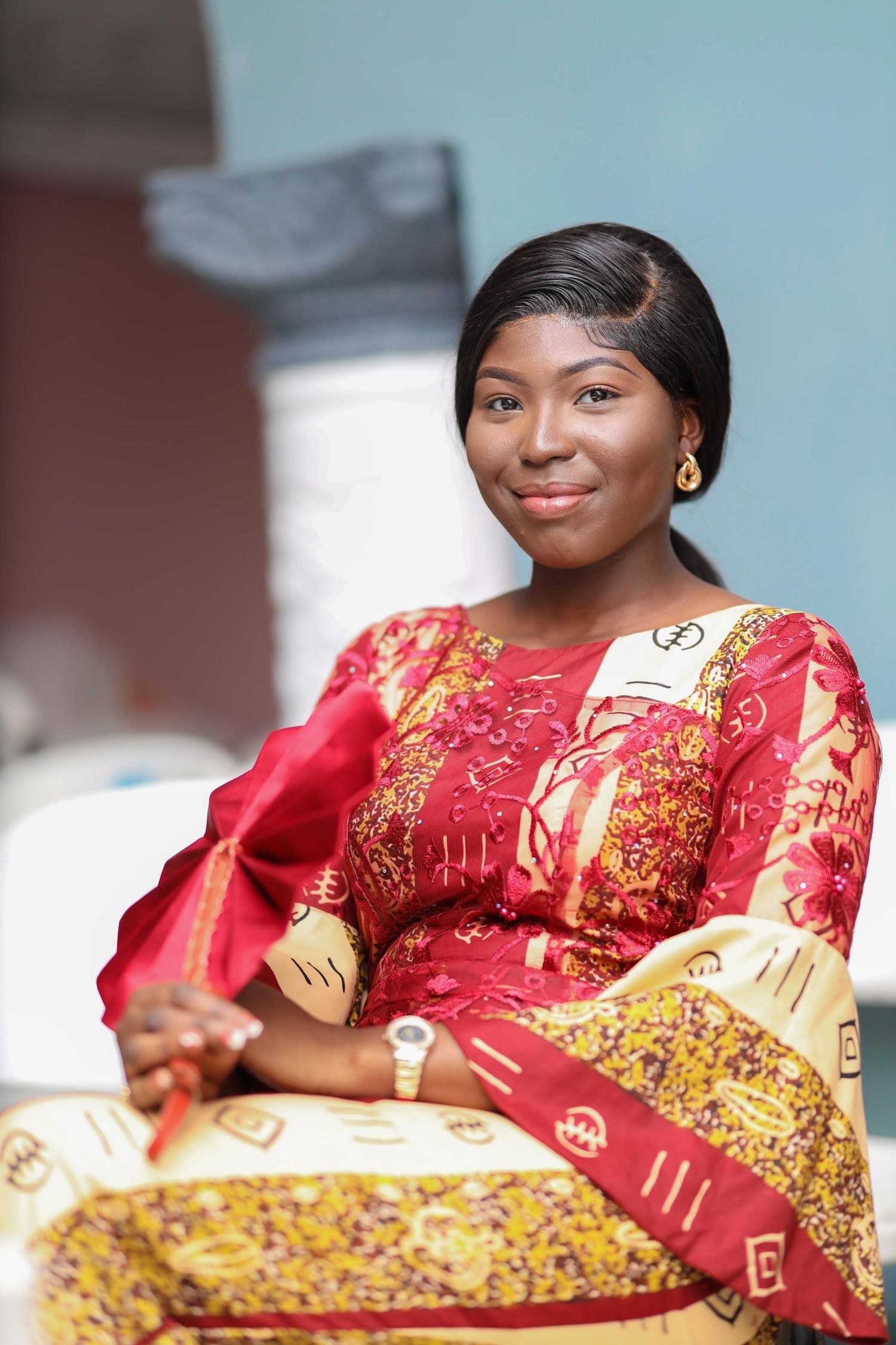 Woman in traditional African dress with red and gold patterns sits elegantly in a bright setting.