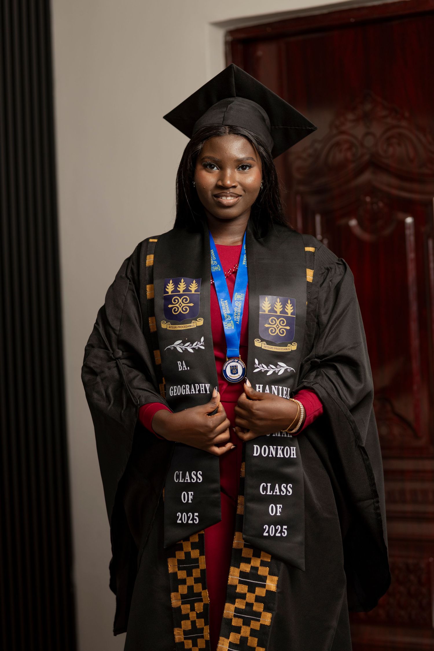 A smiling graduate wearing a black cap and gown with a Class of 2025 stole and medal poses proudly indoors.