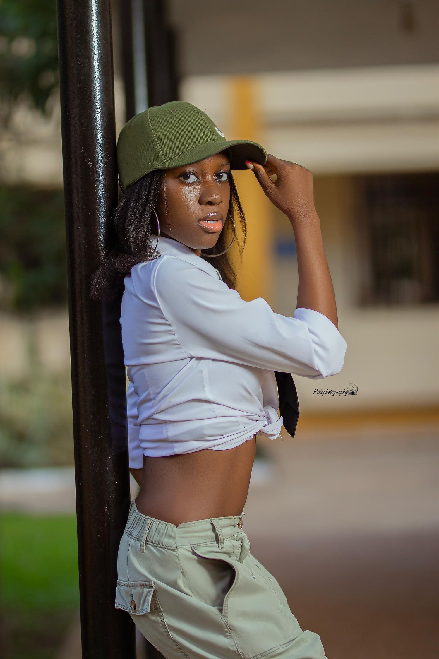 Athletic casual look with green cap, white knotted shirt and cargo pants against pillar.