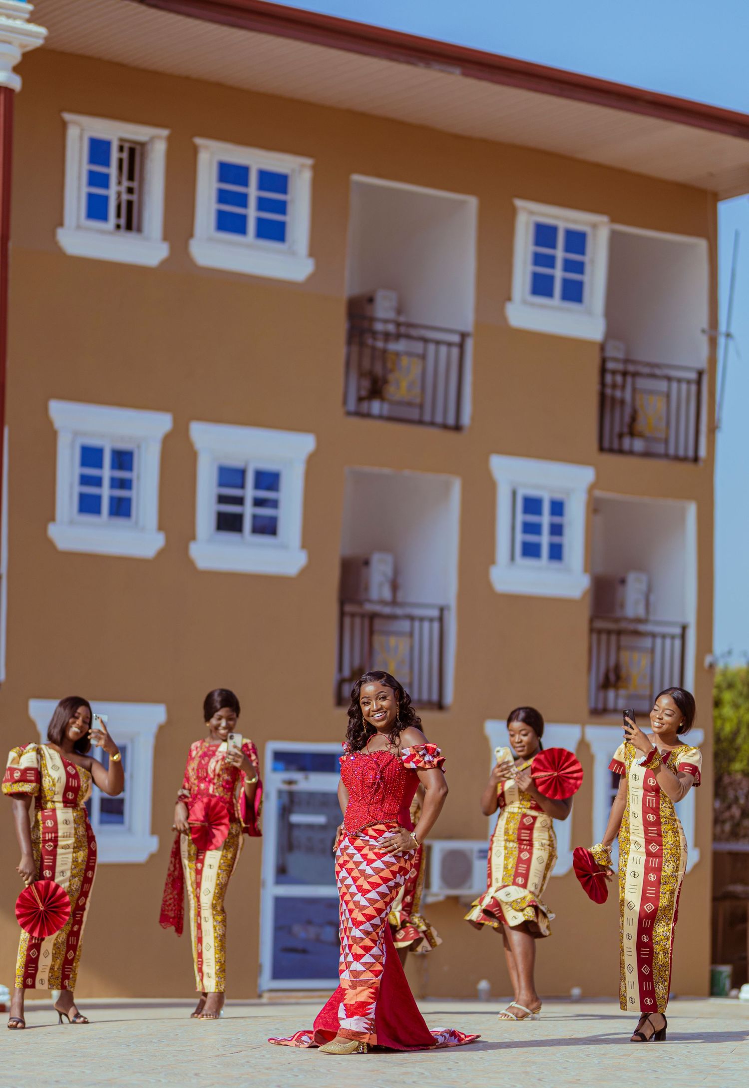 Bridesmaids in red and gold traditional African attire posing in front of a beige building.