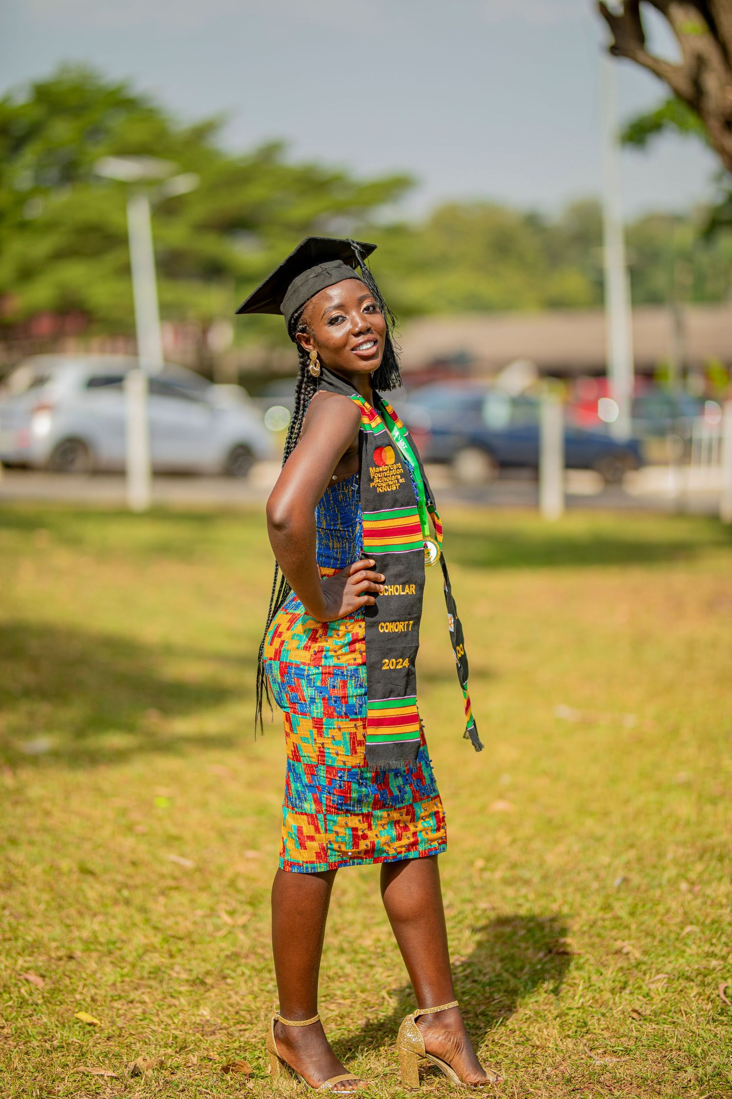 Graduate in colorful patterned dress celebrates joyfully on grassy lawn during outdoor photo session.