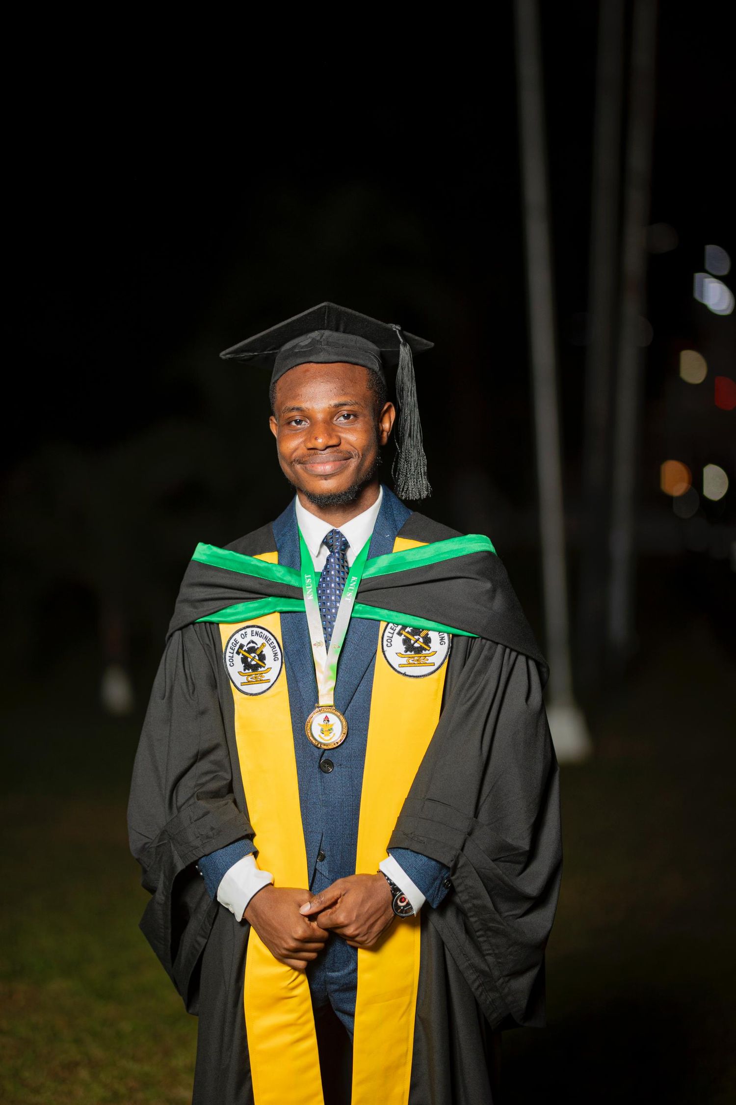 Graduate smiles in traditional black and gold academic regalia with green trim during evening ceremony.