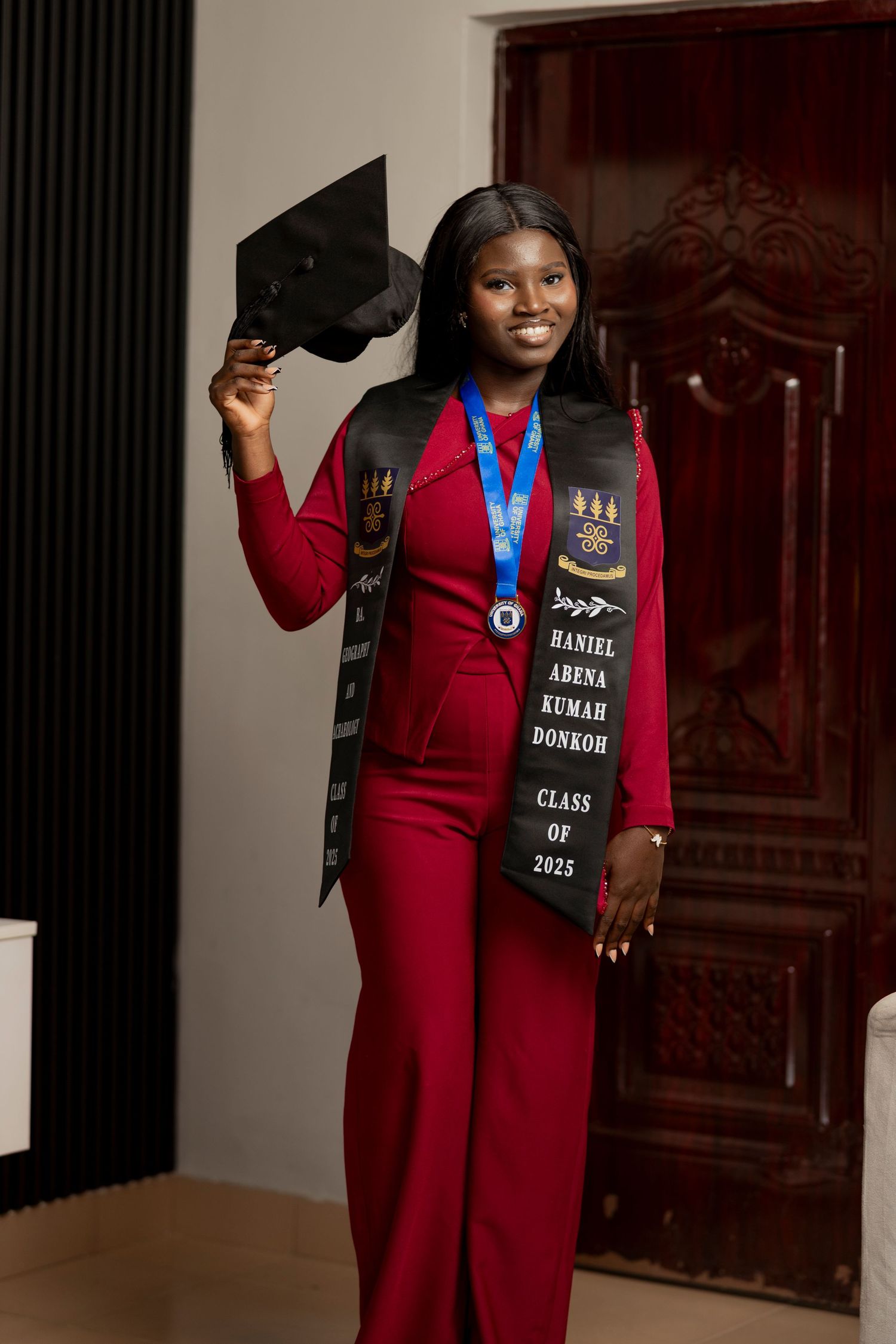 Graduate Haniel Arena Kumah Donkoh poses in red suit with Class of 2025 stole, medal, and mortarboard cap.