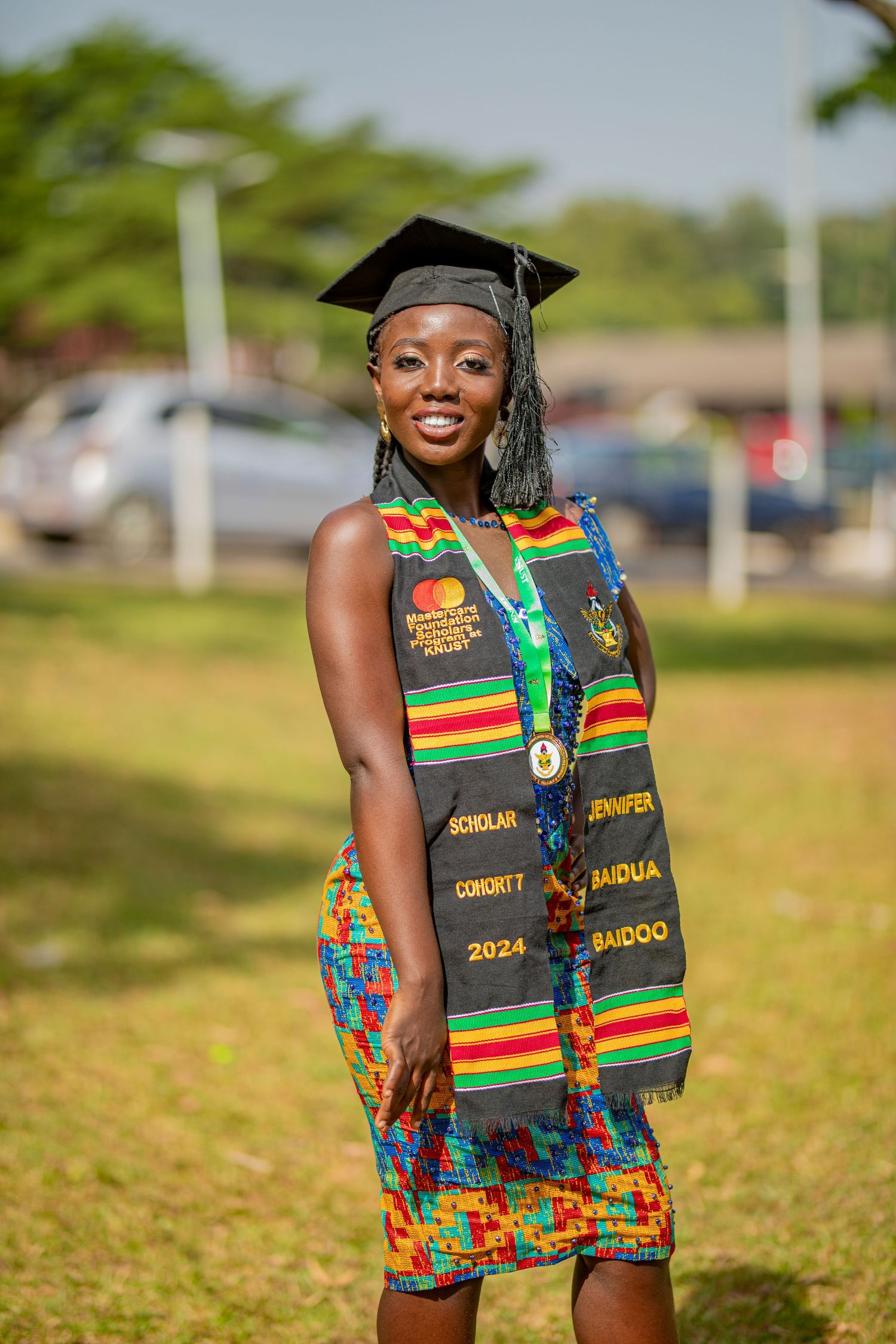 Graduate wearing colorful African kente stole and graduation cap poses outdoors on sunny day.
