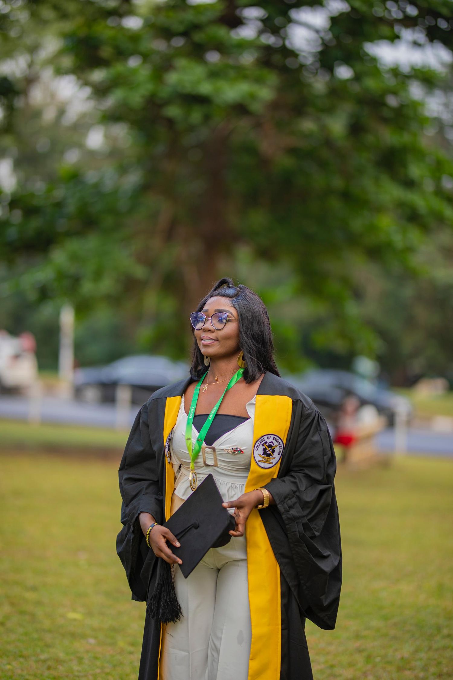 Graduate in academic regalia with yellow stole stands on grass during commencement.