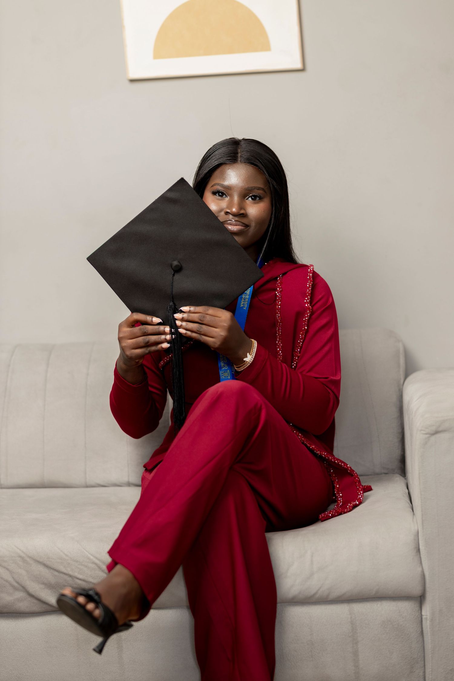 Young Black woman in red suit holding graduation cap while sitting on sofa, celebrating academic achievement.