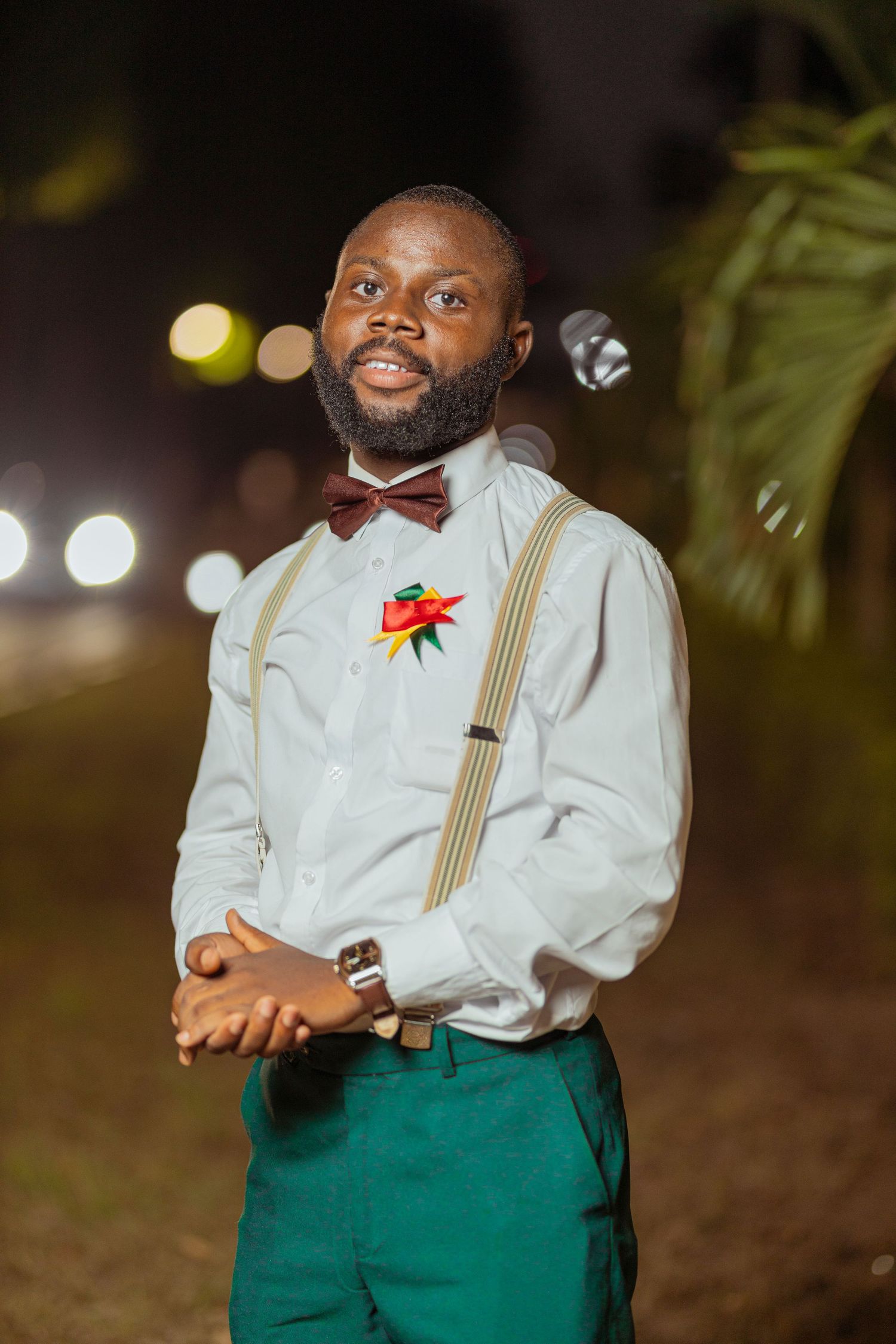 Person in formal attire with suspenders, bowtie and boutonniere poses outdoors at night.