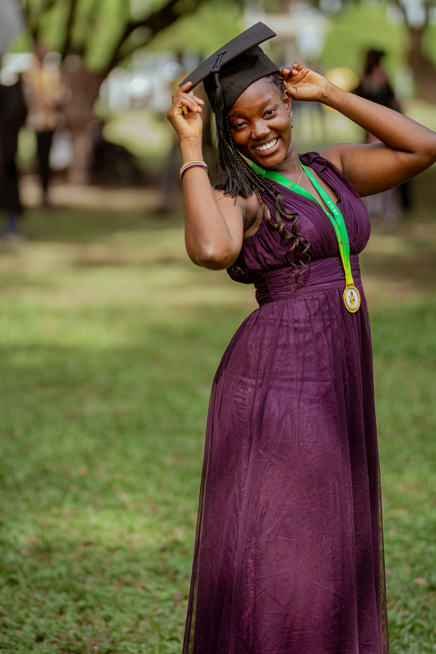Graduate in purple dress and cap holds graduation medal while posing outdoors on grassy lawn.