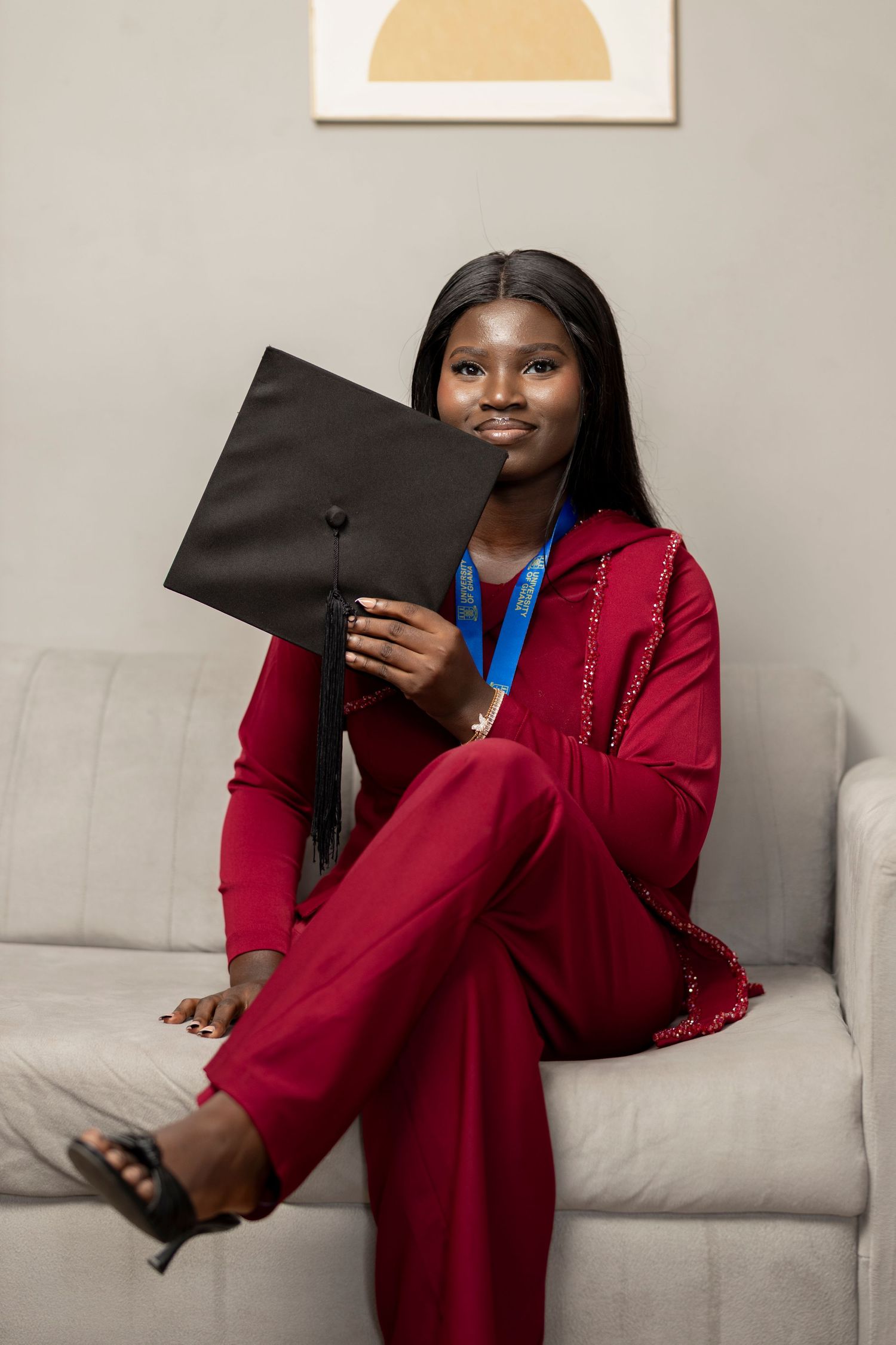 Young woman in red outfit holding graduation cap while sitting on a gray sofa, smiling confidently.