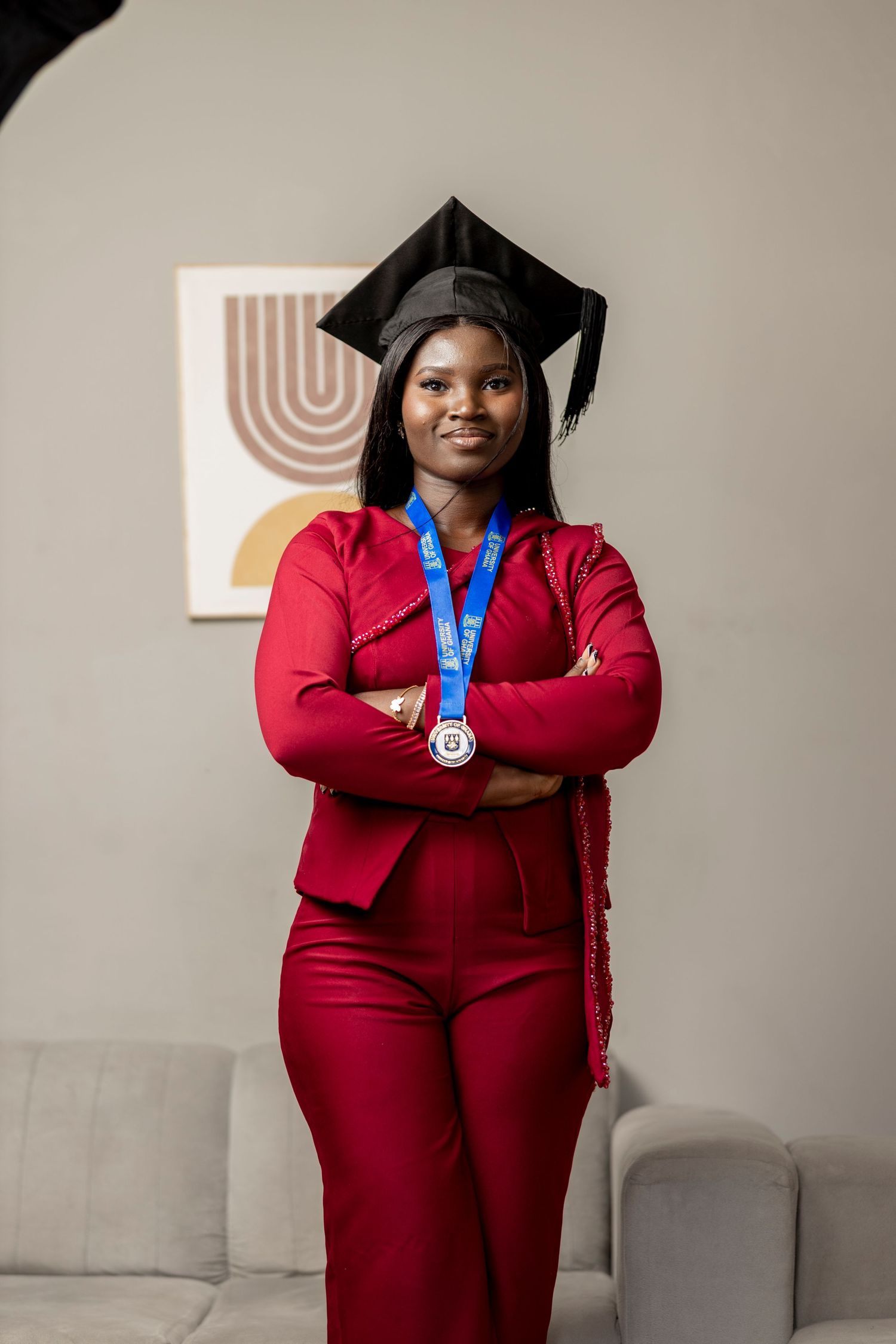 A young woman in a red suit and graduation cap poses confidently with arms crossed, wearing a blue medal.