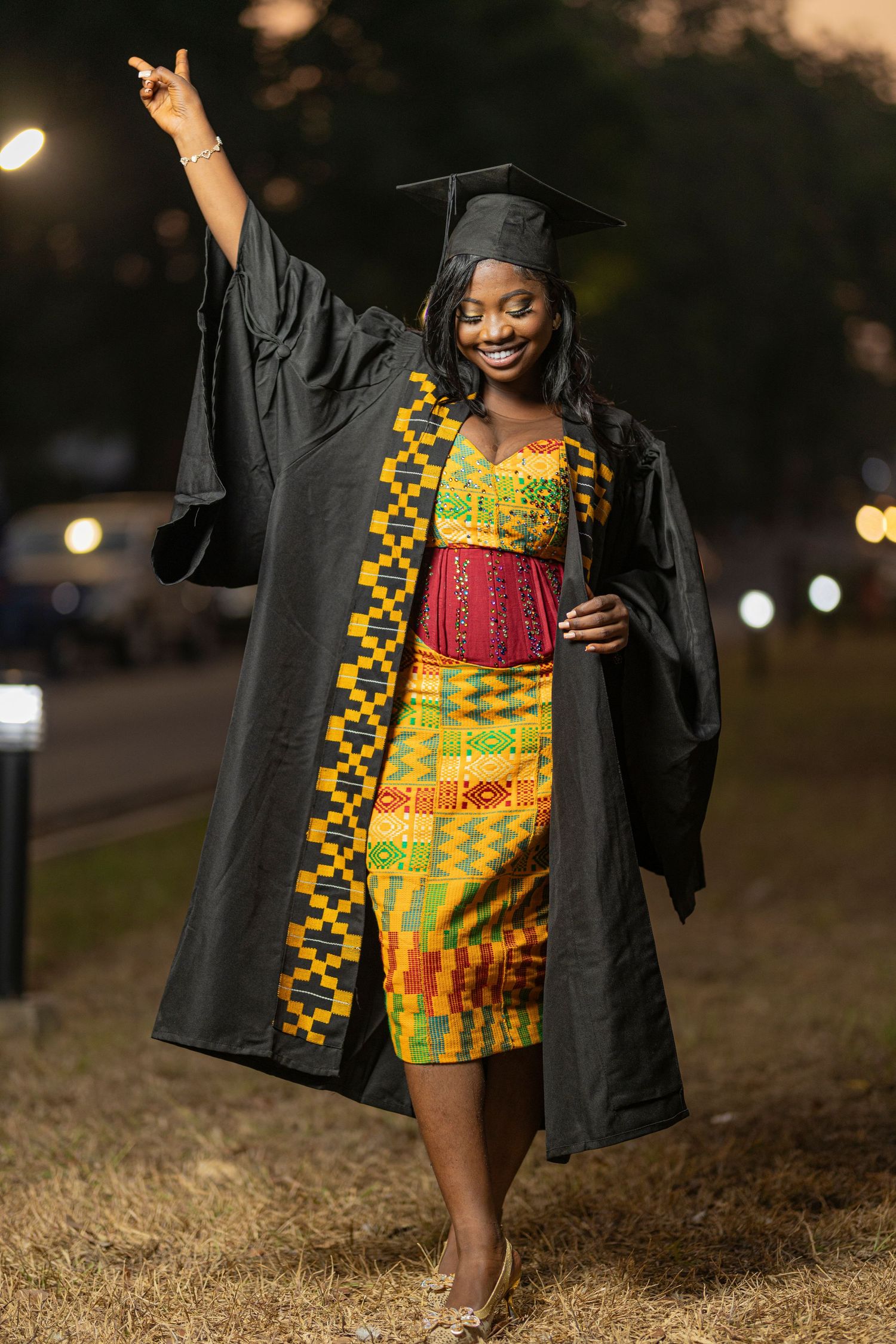 Graduate in black cap and gown with traditional African print dress celebrates at night.