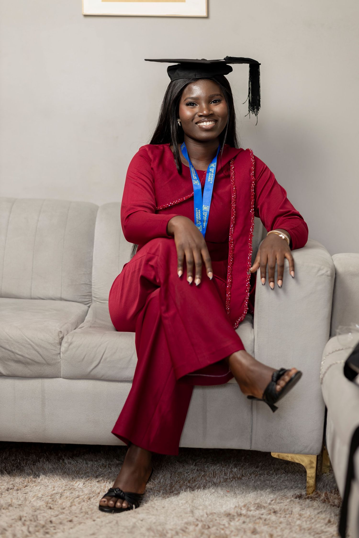 A smiling graduate in red outfit and black cap sits on a gray sofa, wearing a blue medal around her neck.