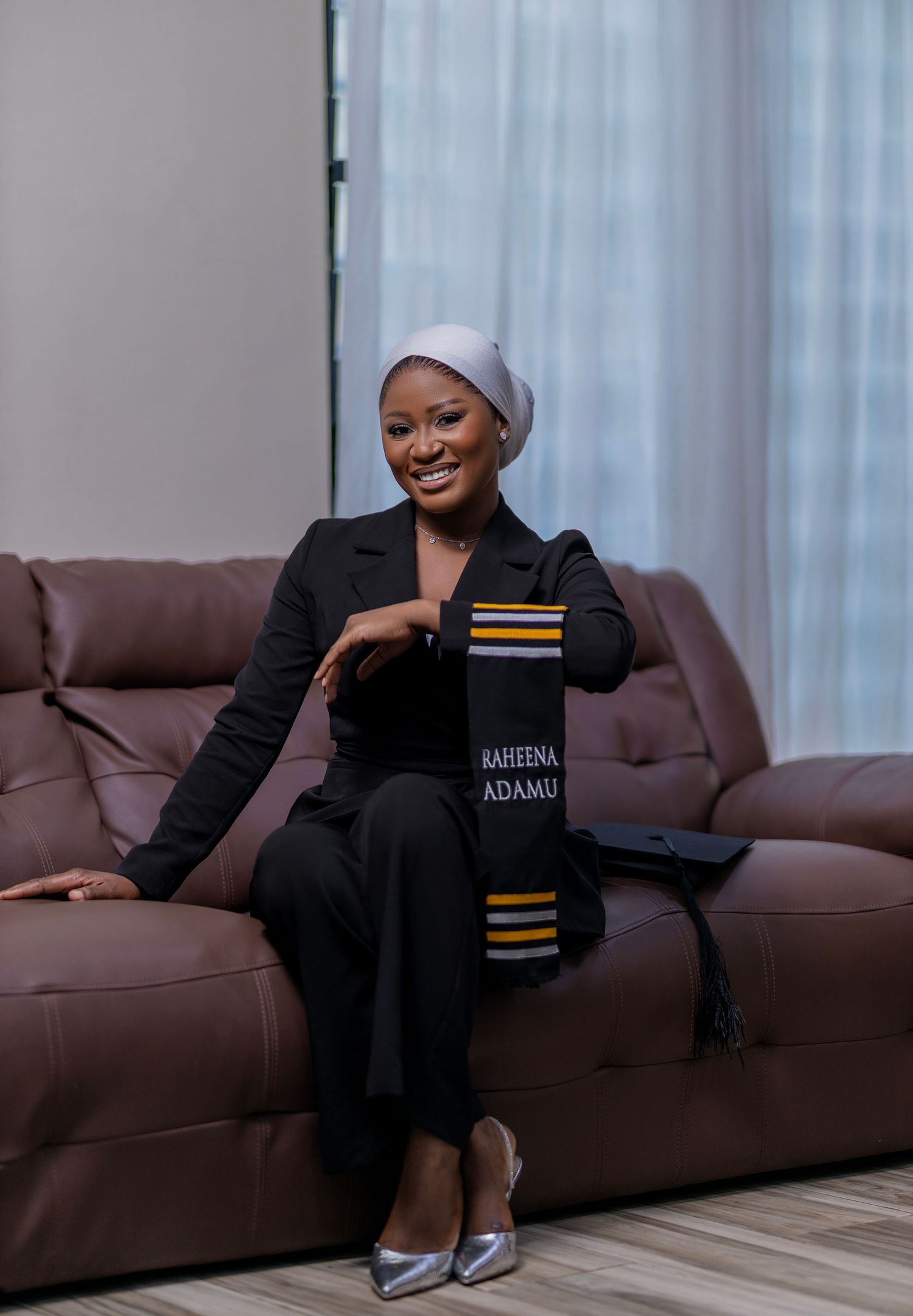 A figure in a black outfit and head covering reclines comfortably on a brown leather couch in a bright room.