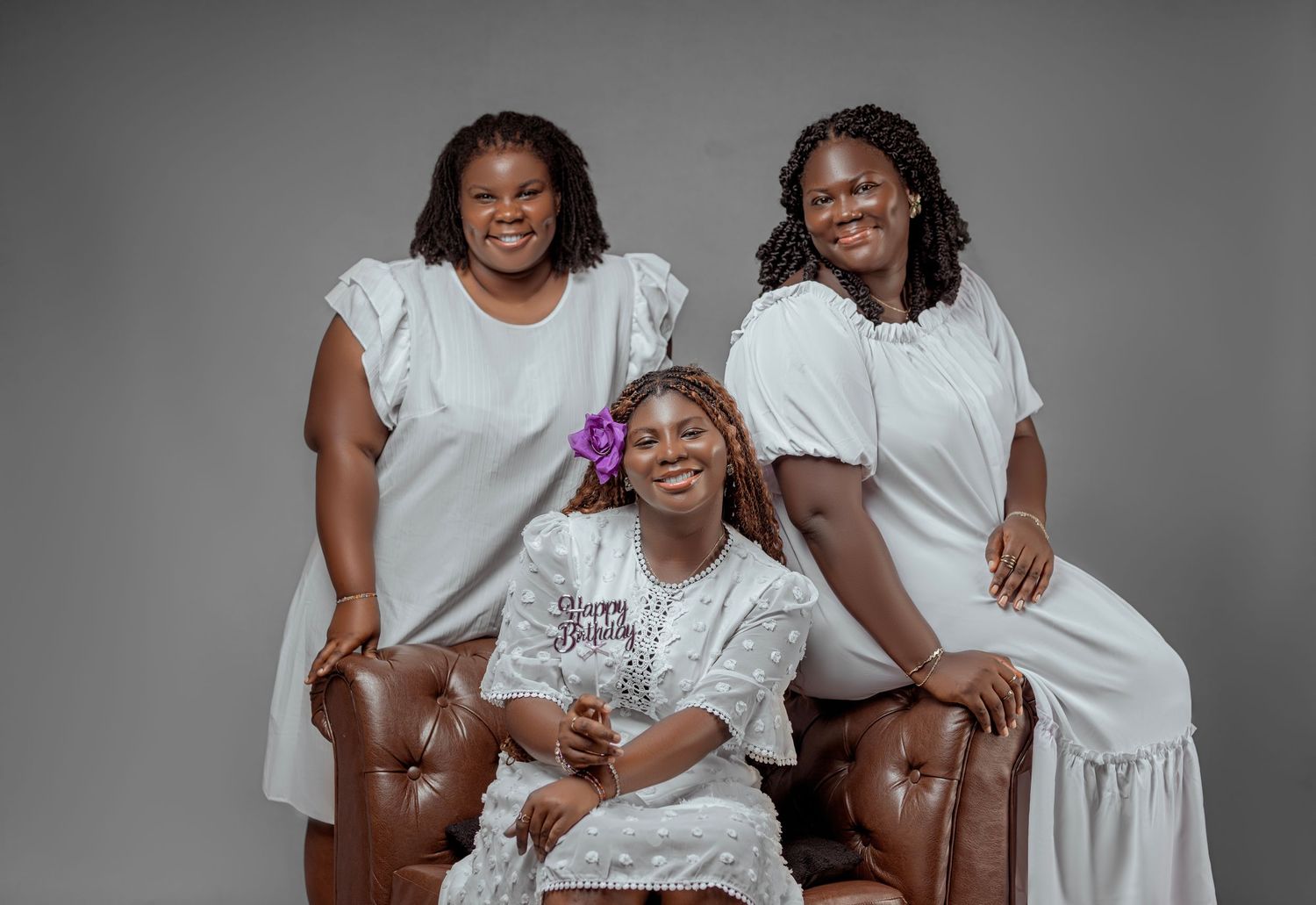 Studio portrait of three women in white dresses posing together against a grey backdrop.