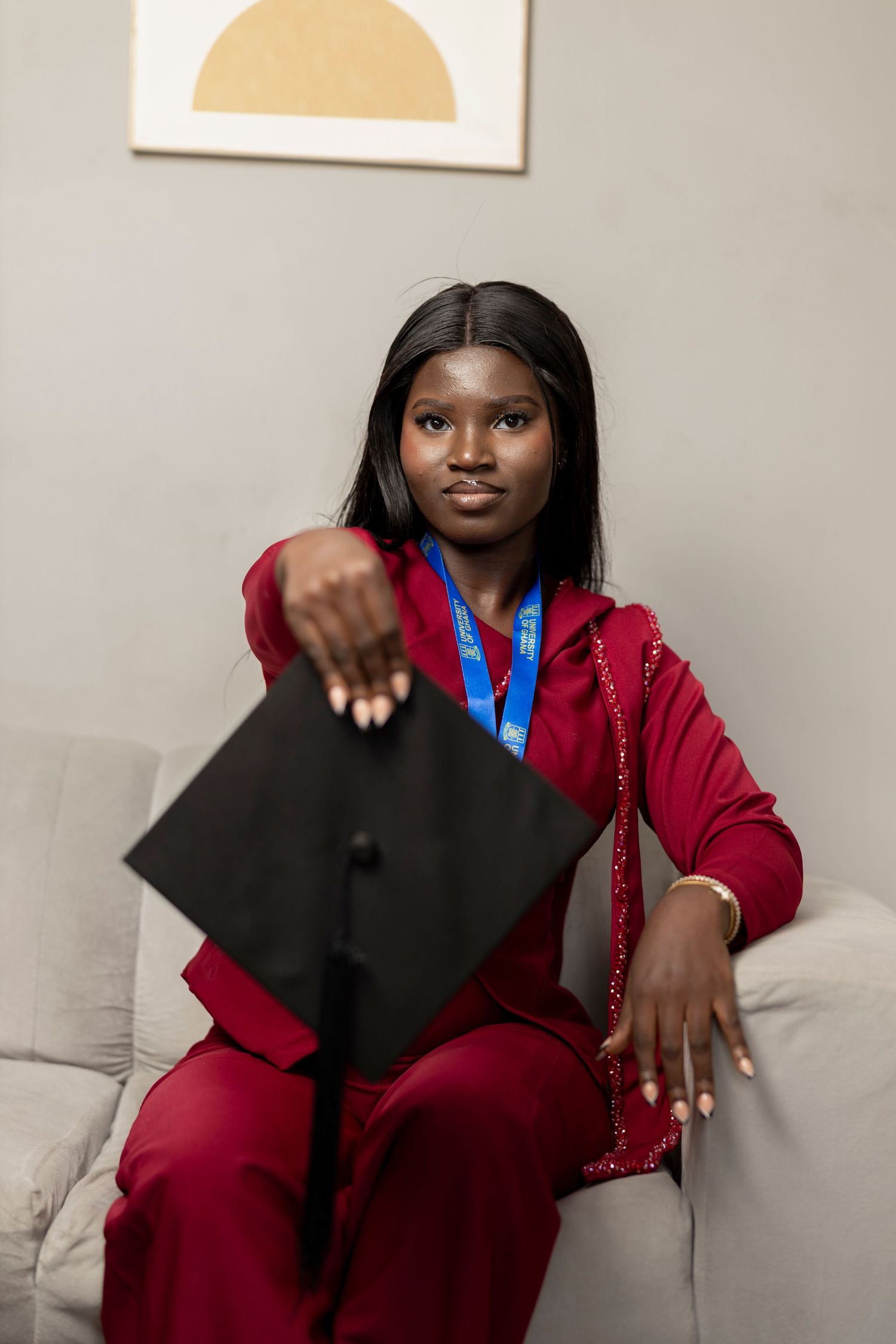 Young Black woman in red suit holding graduation cap with blue medal, sitting on couch in modern room.