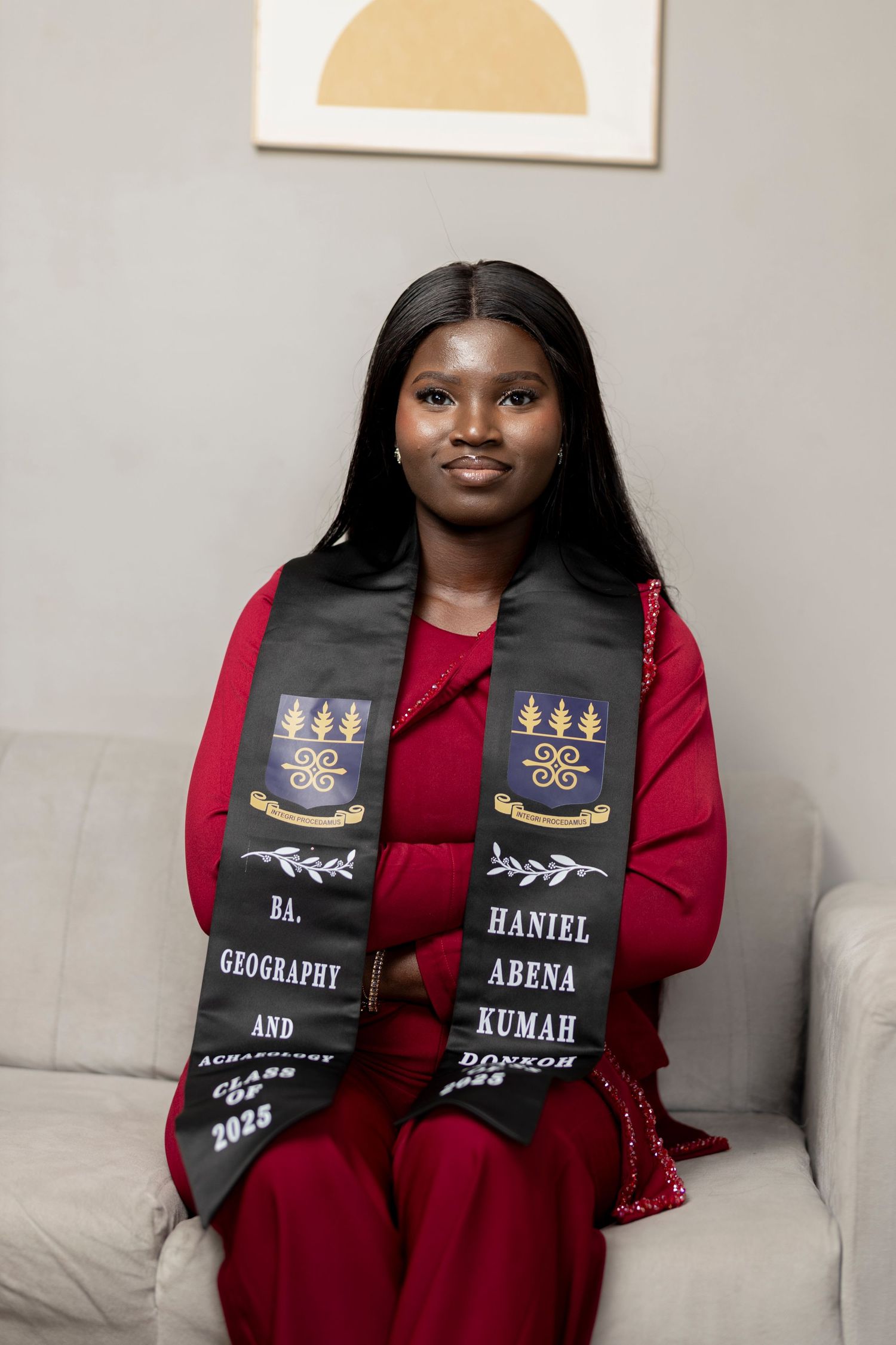 Graduate Haniel Abena Kumah wearing BA Geography graduation stole, Class of 2025, seated in red dress.