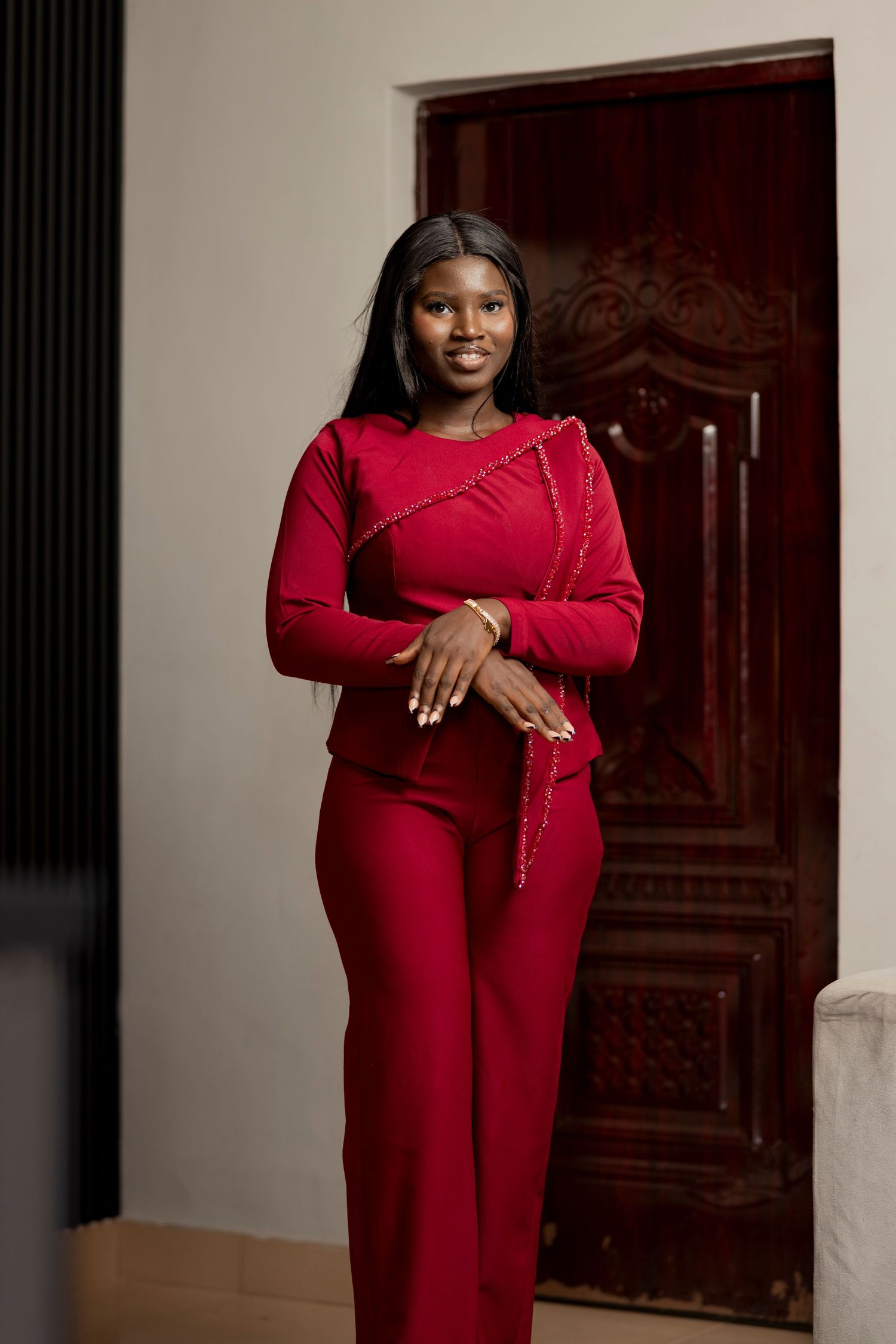 Young woman in elegant red long-sleeve top and wide-leg pants posing confidently near ornate wooden door.