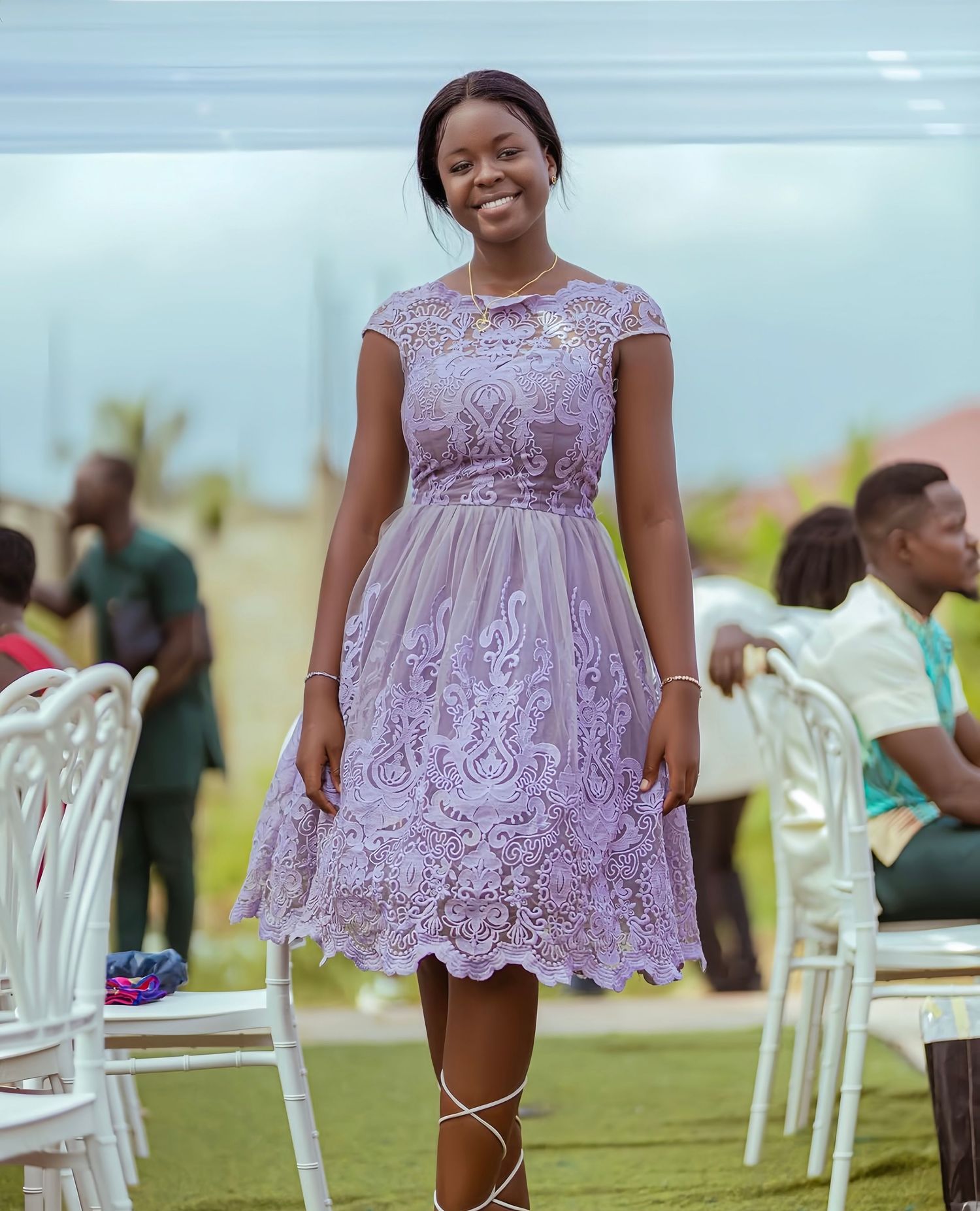 A person wearing a lovely lavender lace dress stands outdoors at an elegant event with white chairs on a lawn.