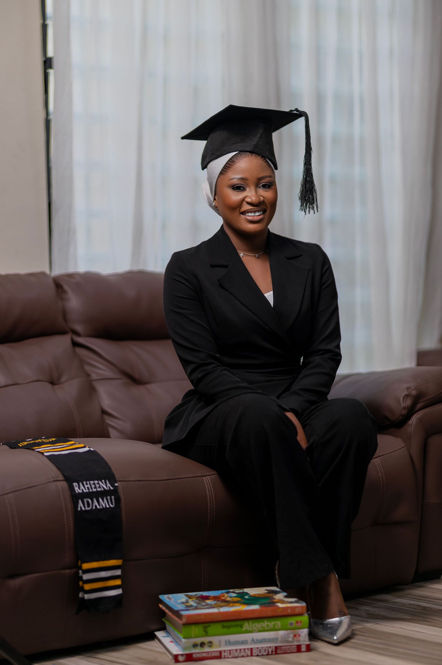 Graduate in black academic regalia poses with books and personalized name sign on brown couch for celebration photos.