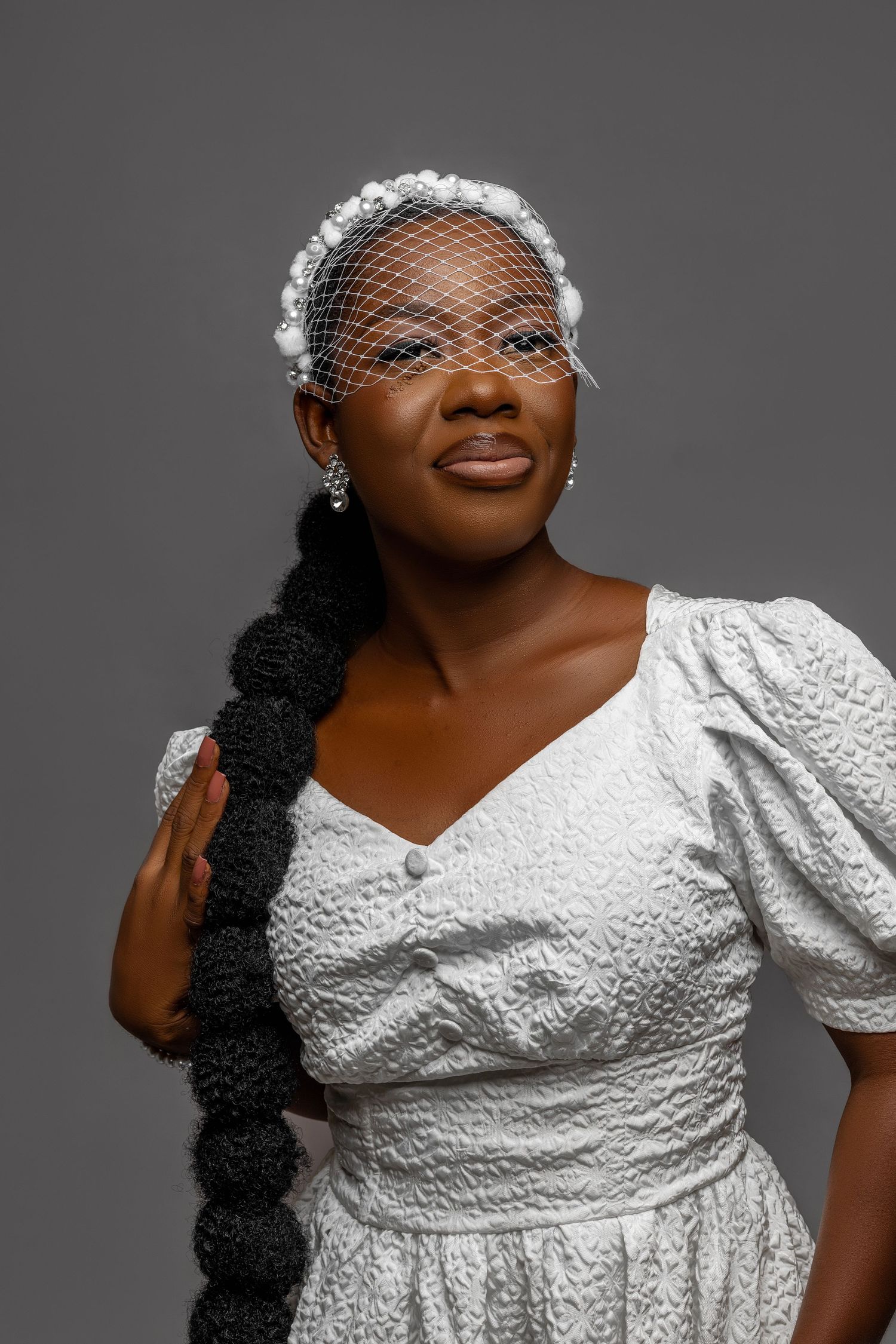A person wearing a white vintage-style dress with lace detail and a small netted headpiece poses against a gray background.