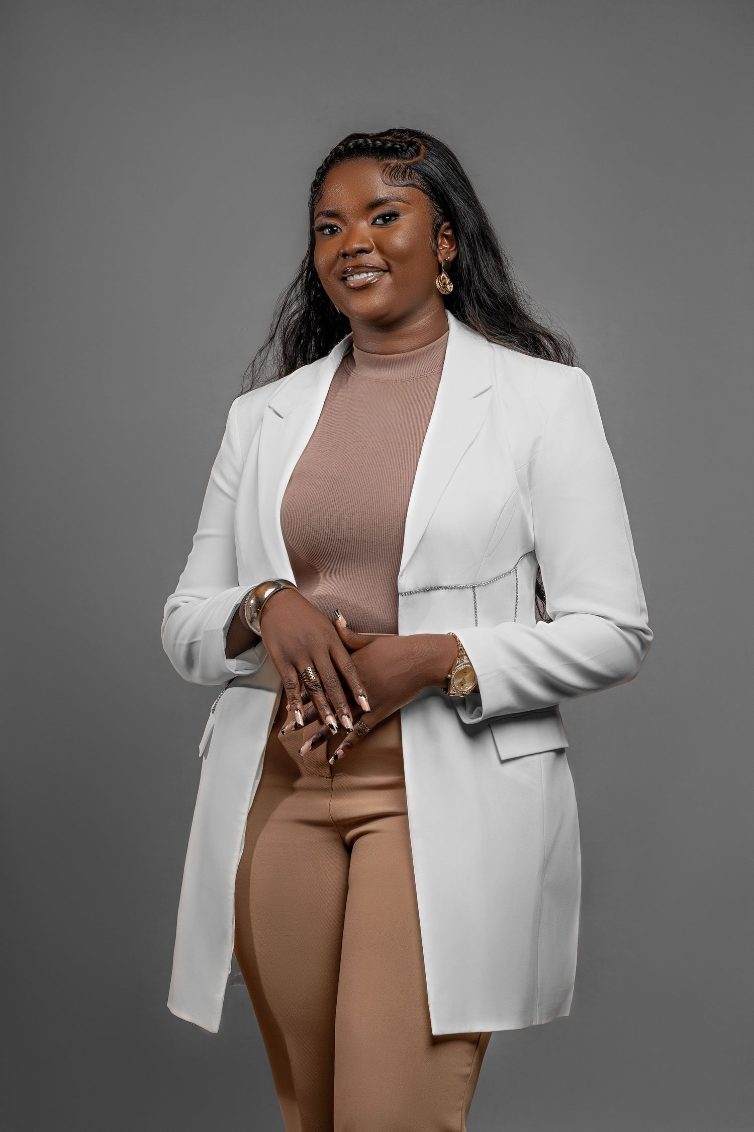 Professional studio portrait showing confident pose in white blazer and tan outfit against gray background.