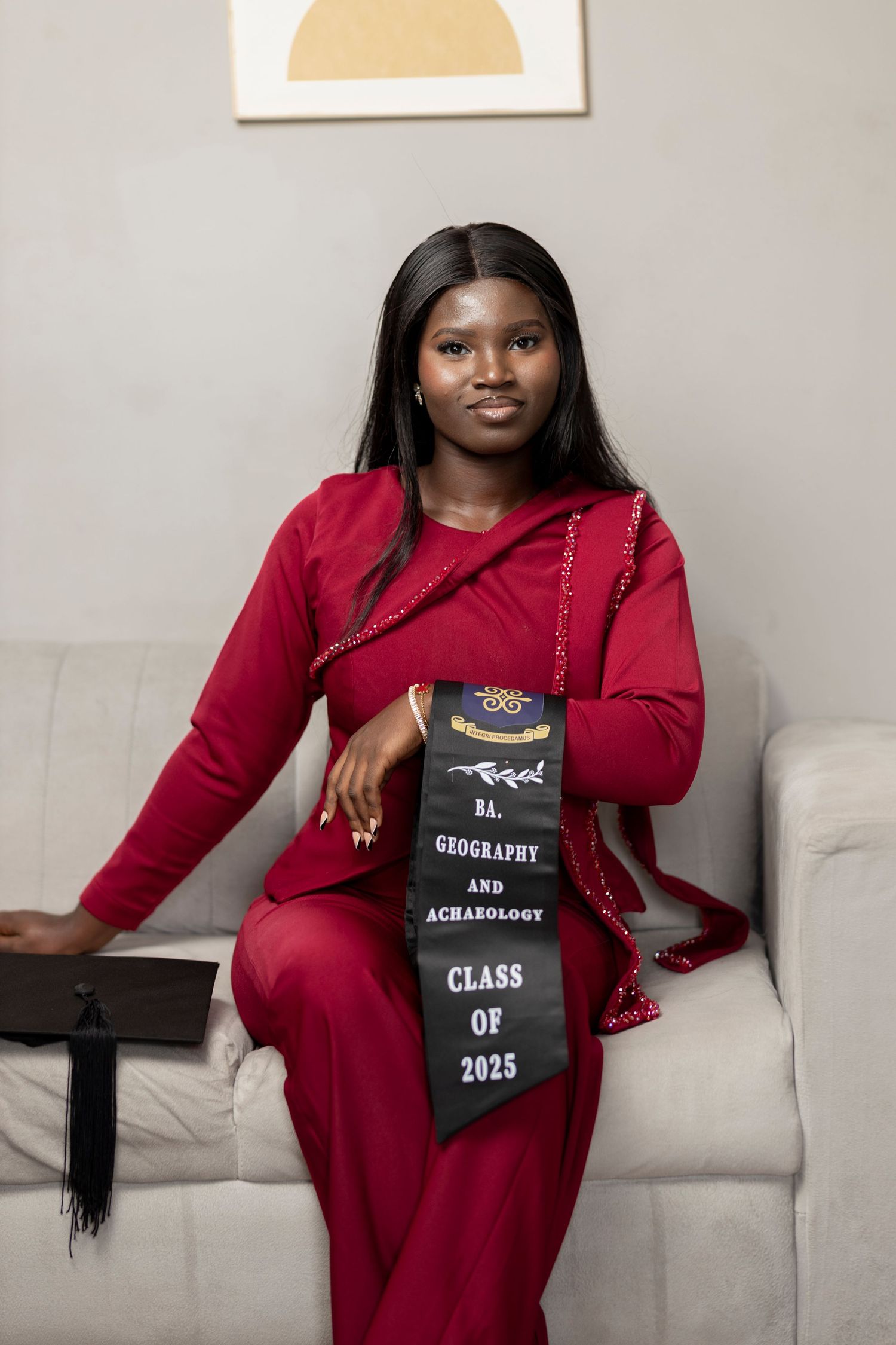 Young woman in red dress wearing BA Geography and Archaeology Class of 2025 graduation sash seated on white sofa.