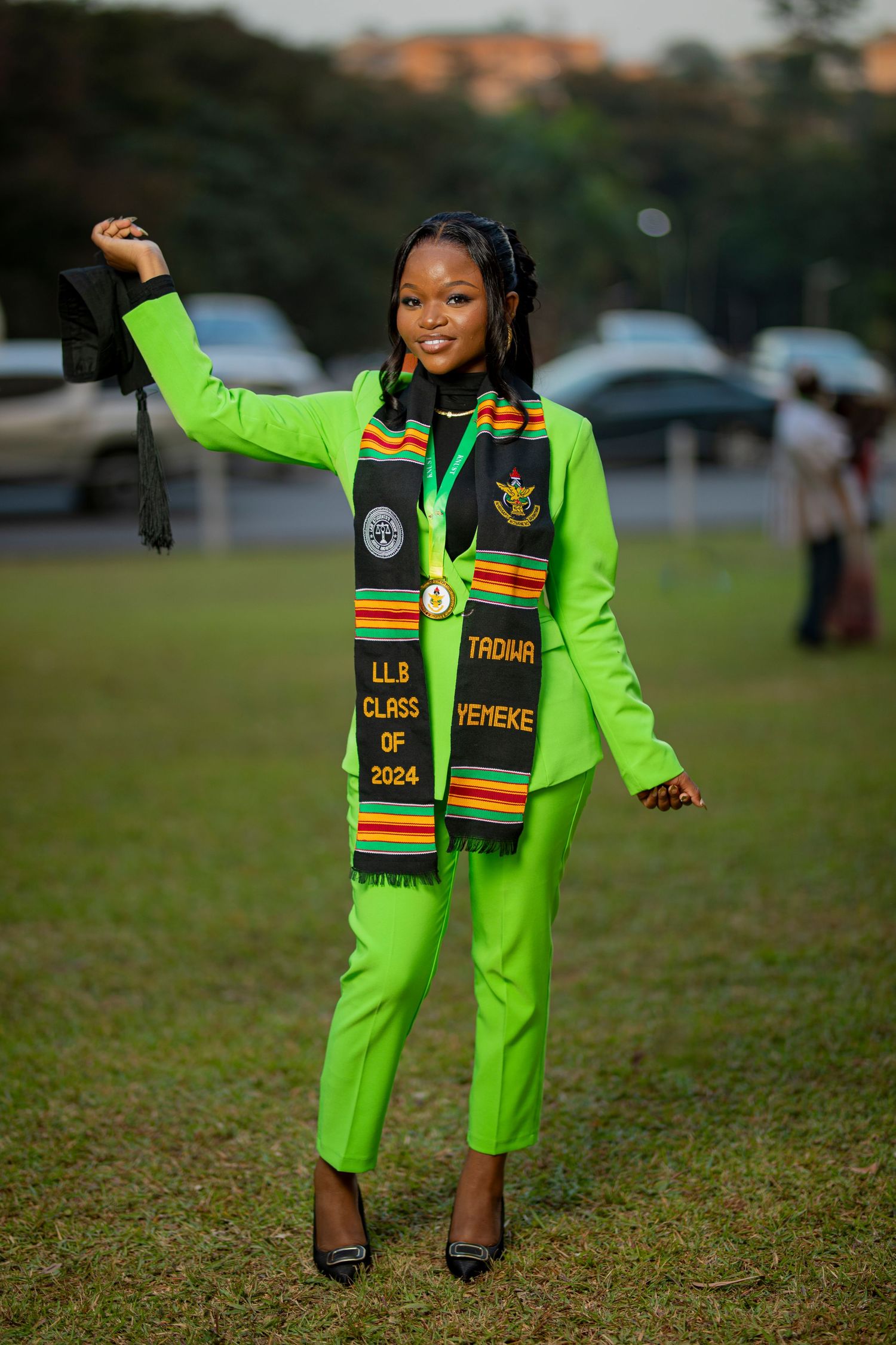 Graduate wearing vibrant neon green outfit and class of 2024 graduation stole poses joyfully on grass field.