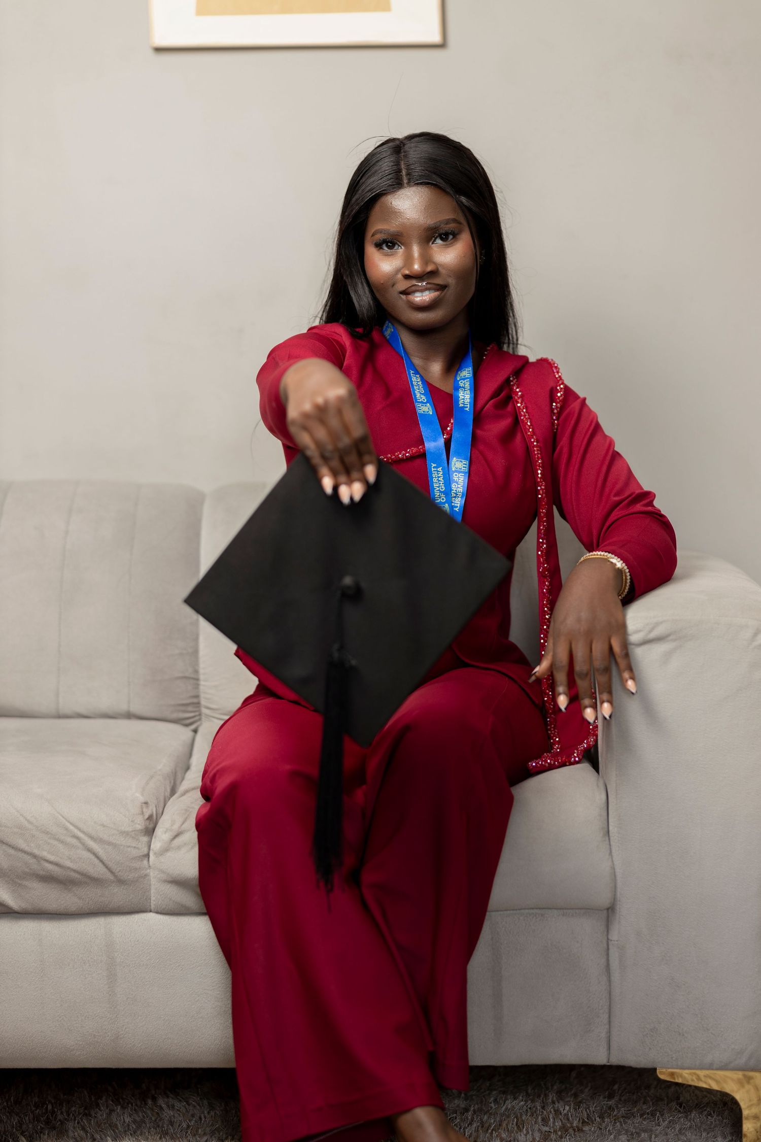 Young woman in red outfit holding graduation cap while wearing blue lanyard, seated on gray sofa.