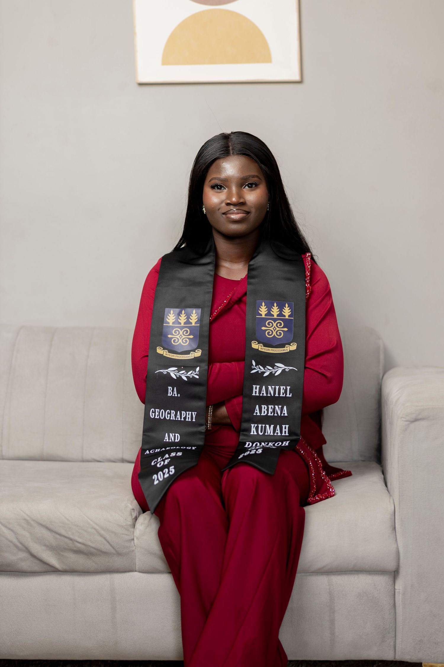 Young woman in red outfit wearing graduation stole reading Geography, seated on white couch for graduation photo.