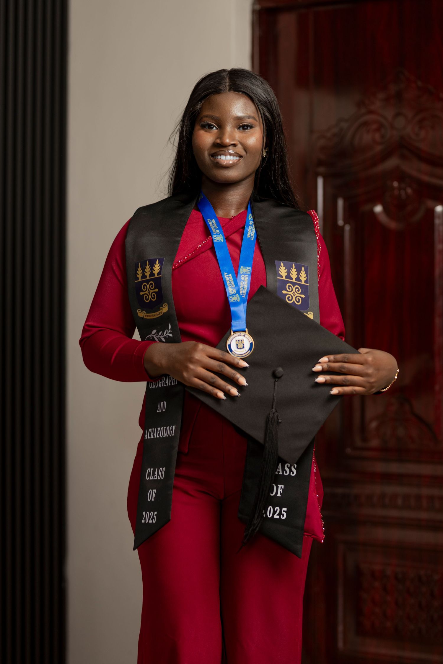 Smiling graduate in red outfit holds cap, wearing blue medal and Class of 2025 stole at graduation portrait.