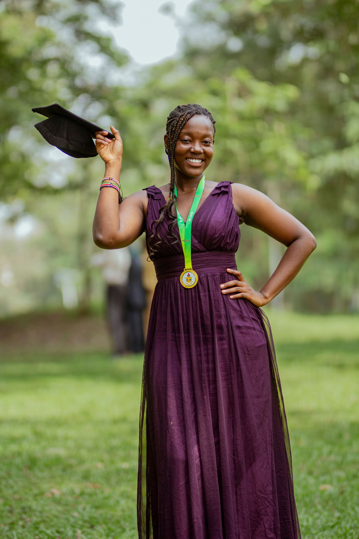 A person in a purple dress poses joyfully in a green outdoor setting while holding a graduation cap and medal.