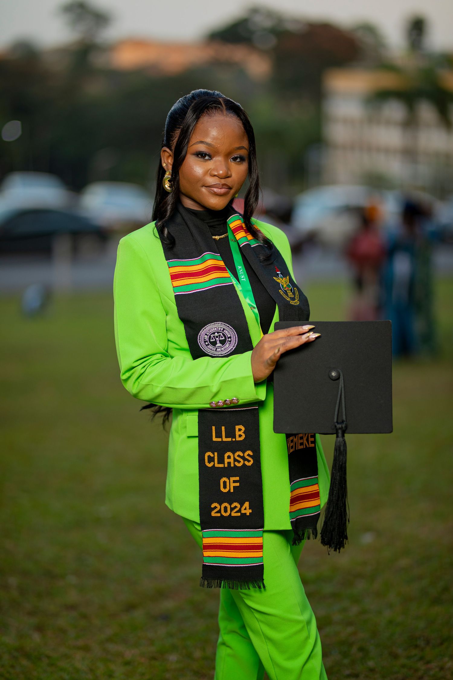Graduate in bright neon green academic regalia and kente stole poses with diploma during outdoor commencement ceremony.