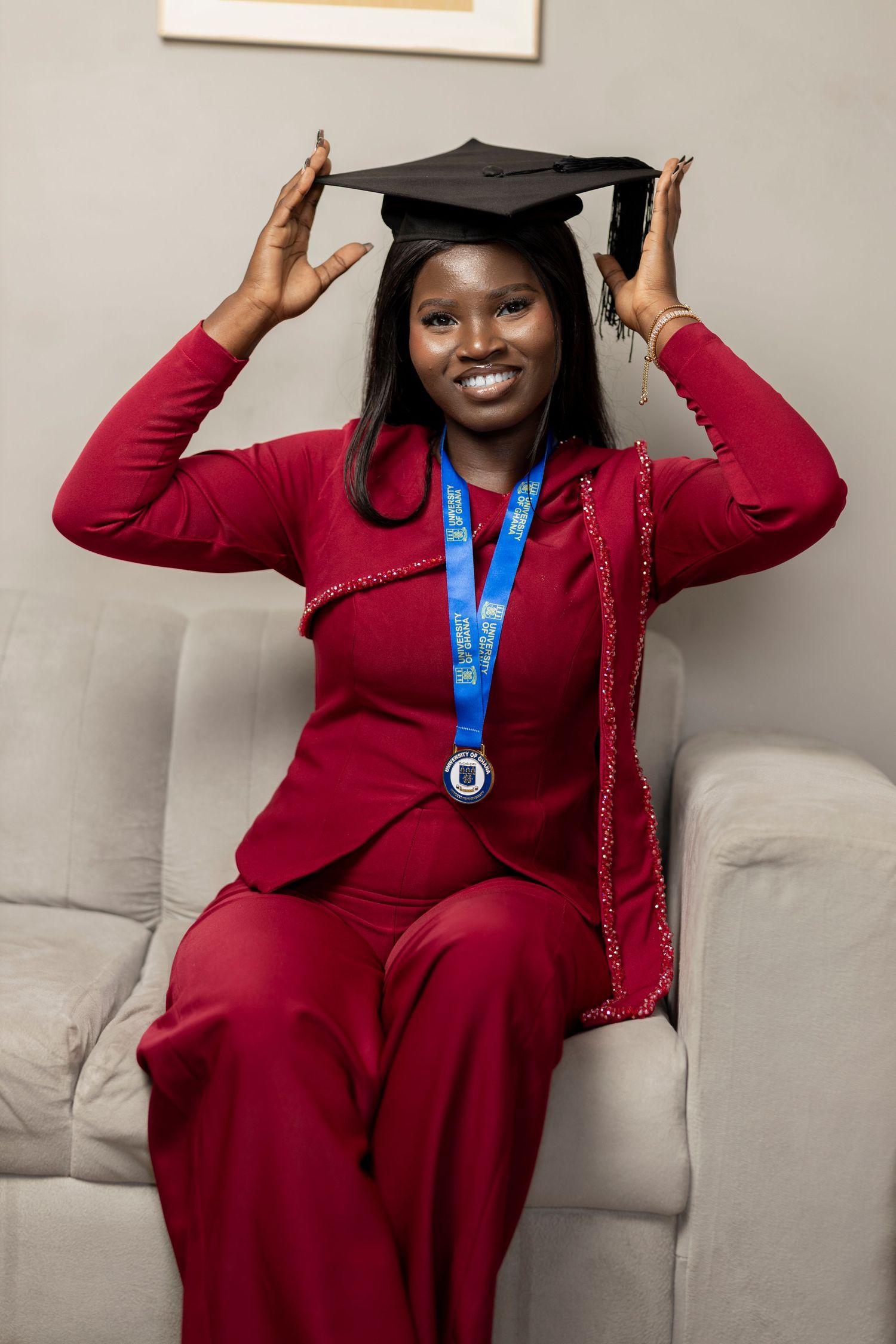 Smiling graduate in red suit wearing mortarboard cap and blue medal, sitting on white sofa celebrating achievement.