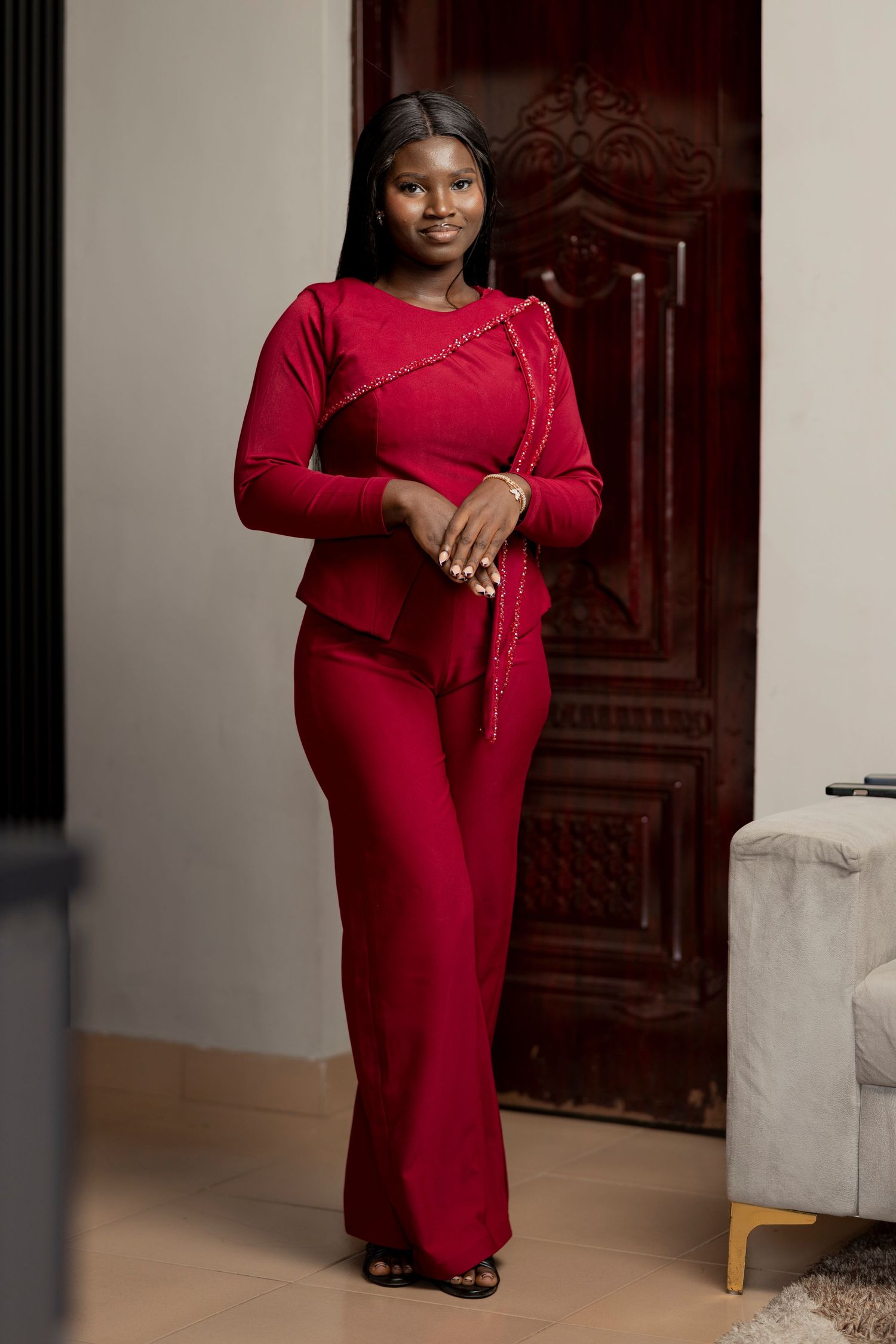 Woman in elegant red two-piece outfit with gold trim detail, posing indoors near a wooden door.